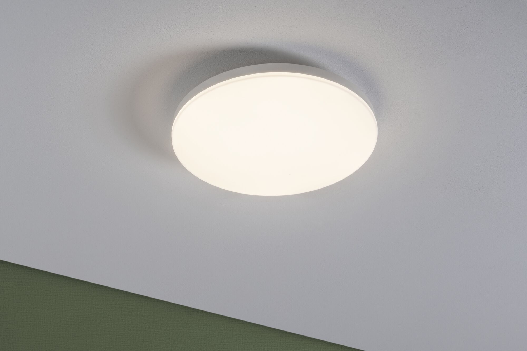 Eine runde, weiße Deckenlampe leuchtet an einer weißen Decke mit einem grünen Wandstreifen.