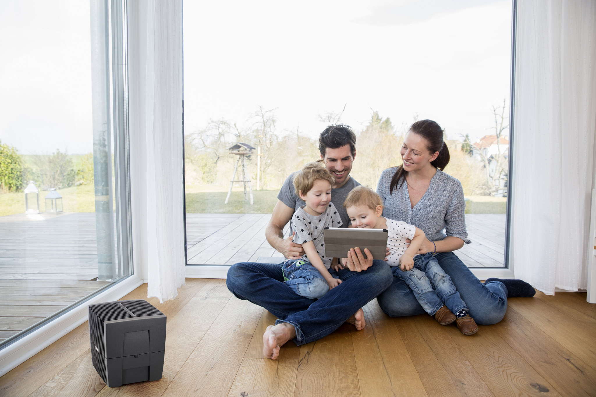 Eine glückliche Familie sitzt auf einem Holzfußboden vor einem großen Fenster und nutzt gemeinsam ein Tablet.