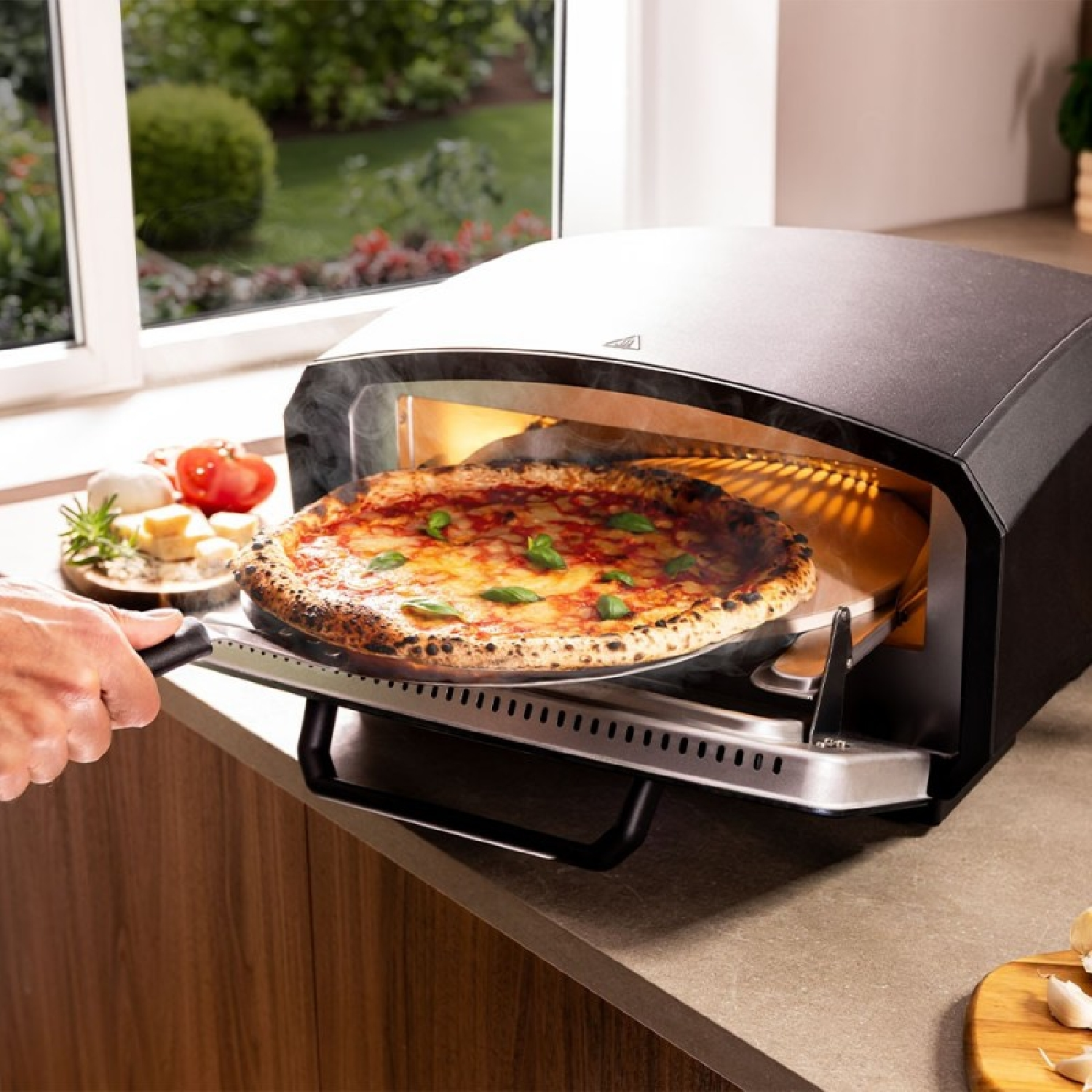 Una mano introduce una pizza recién horneada con albahaca en una pala dentro de un horno para pizza de sobremesa negro.