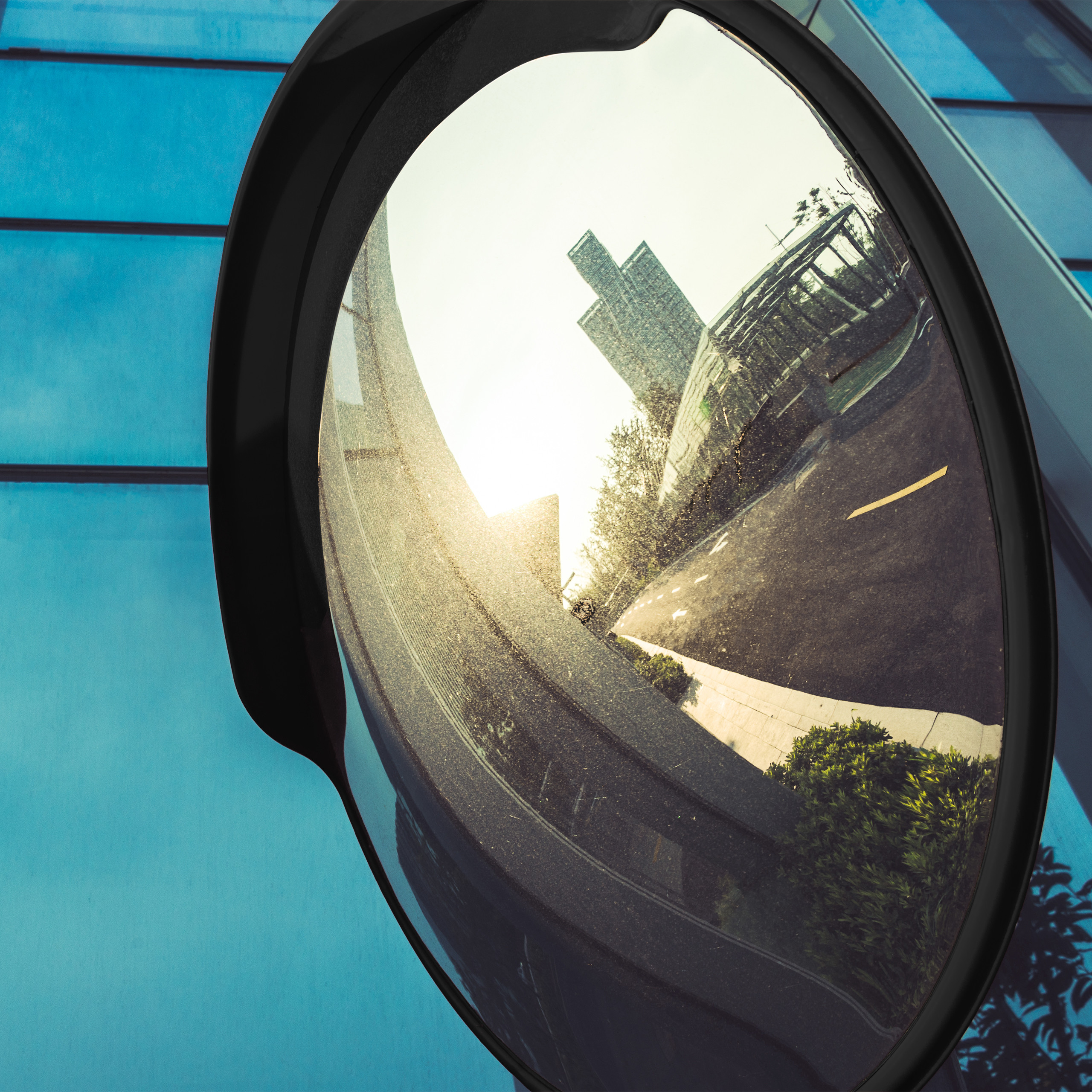 Ein gewölbter Verkehrsspiegel zeigt die Reflexion einer sonnigen Straße mit Gebäuden und Bäumen vor einer blauen Fassade.