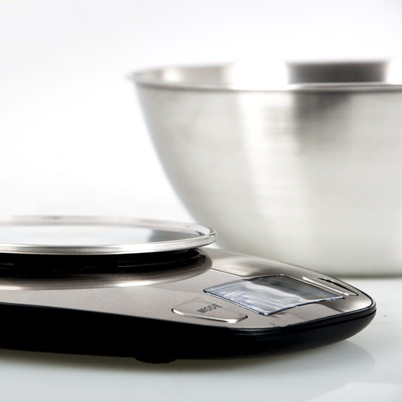 Una balanza de cocina plateada sobre una superficie blanca, con un bol de acero inoxidable detrás.