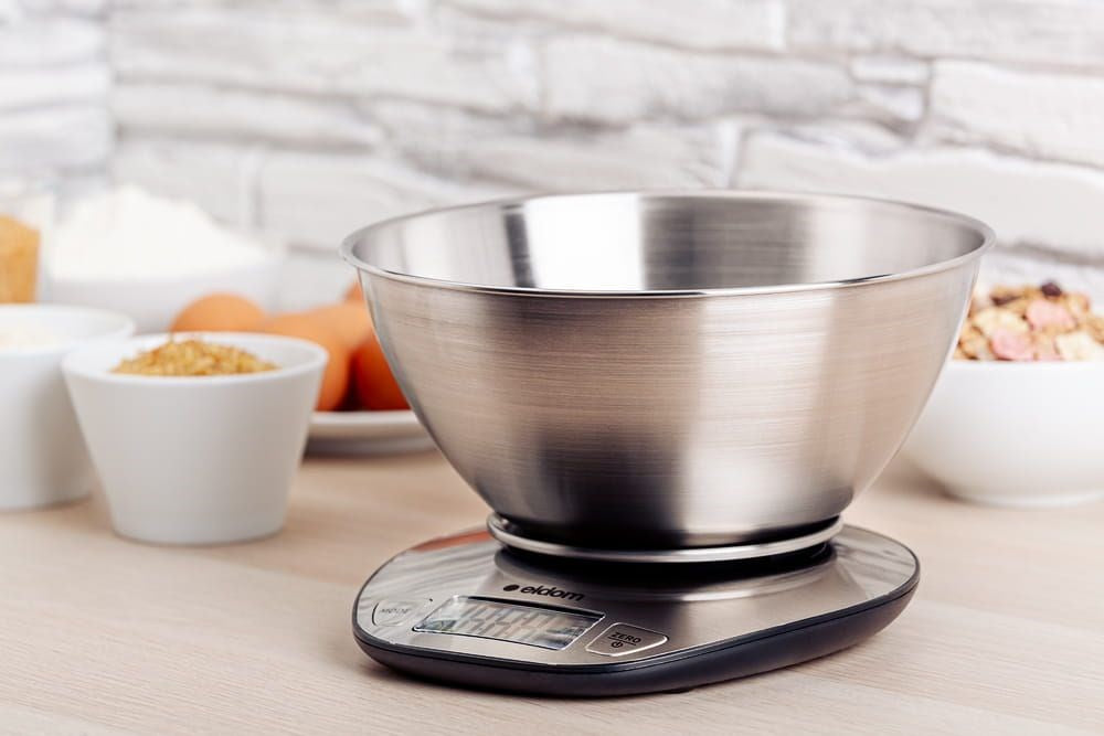 Un bol de mezcla de acero inoxidable sobre una balanza de cocina digital. Ingredientes de repostería al fondo.