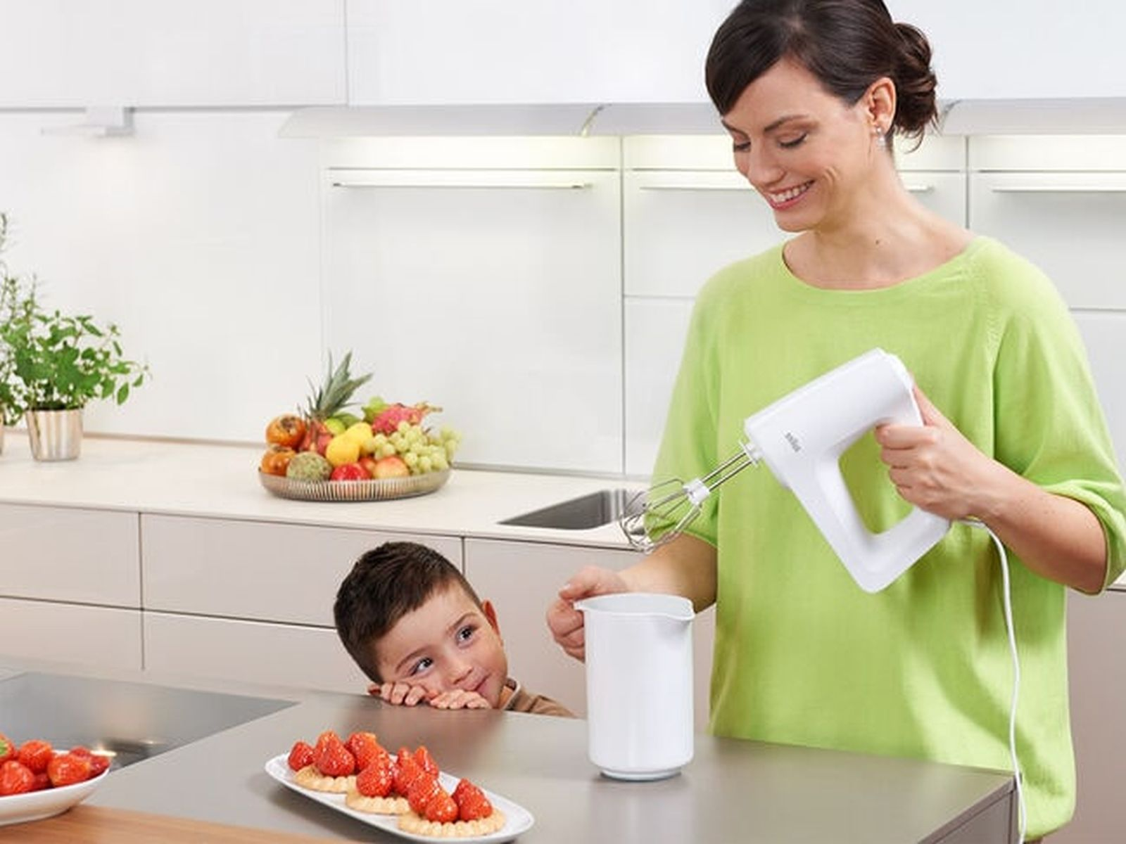 Eine Frau in grünem Oberteil nutzt einen Handmixer, während ein Kind zusieht. Auf dem Tisch stehen Erdbeertörtchen.