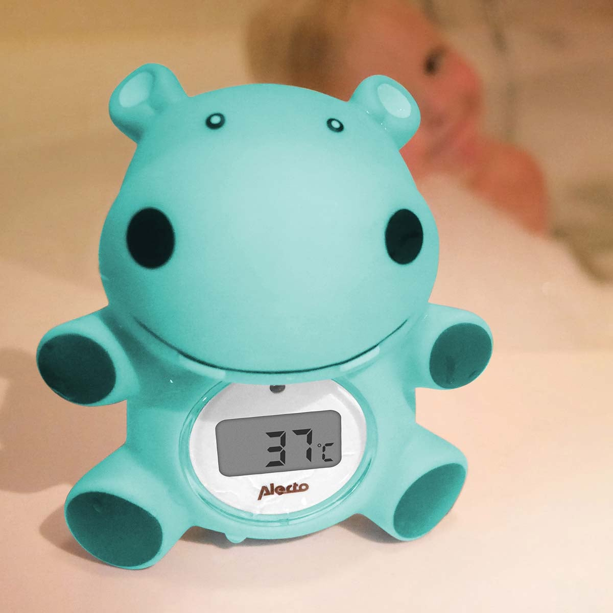 Un termómetro de baño de hipopótamo turquesa muestra 37 grados en una pantalla digital. Un bebé borroso se ve al fondo.