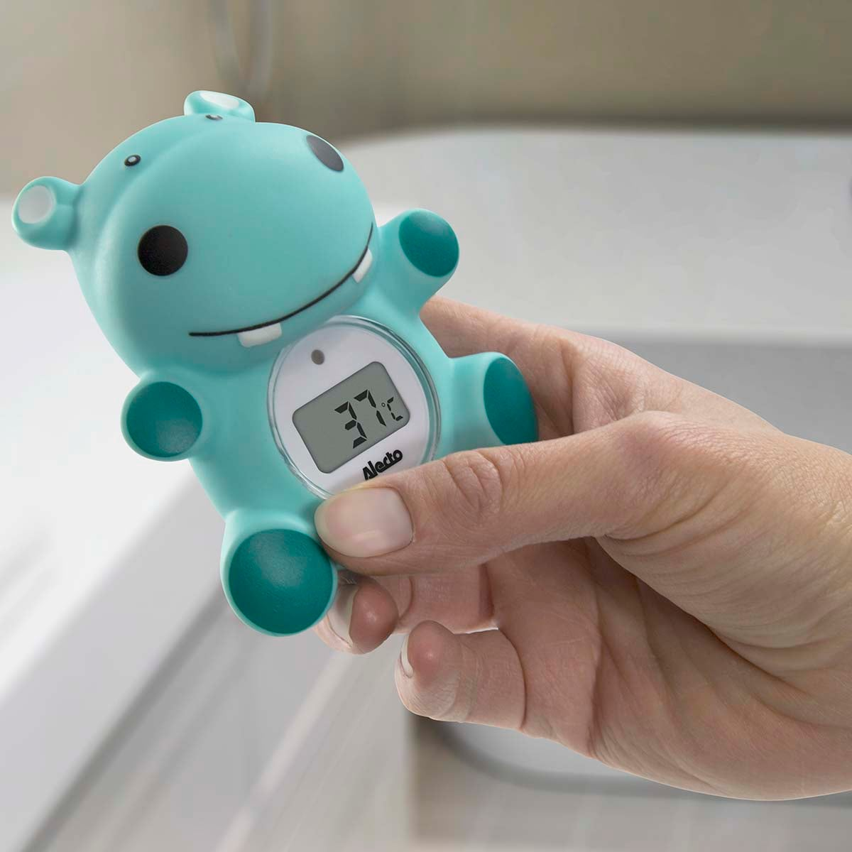 Una mano sostiene un termómetro de baño de hipopótamo turquesa que muestra 37 grados.