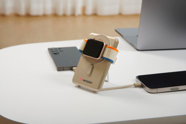 Eine goldene Verbatim Powerbank auf einem weißen Tisch lädt eine Smartwatch und zwei Smartphones, mit Laptop im Hintergrund.