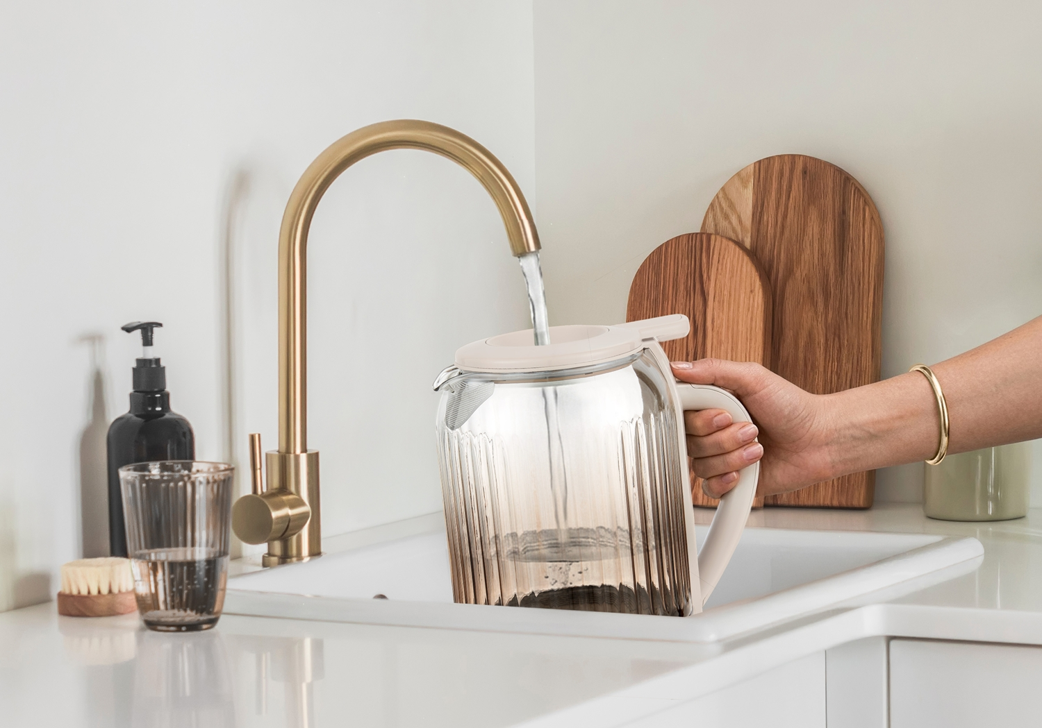 Una mano riempie una caraffa di vetro scanalata con acqua da un rubinetto dorato sopra un lavandino.