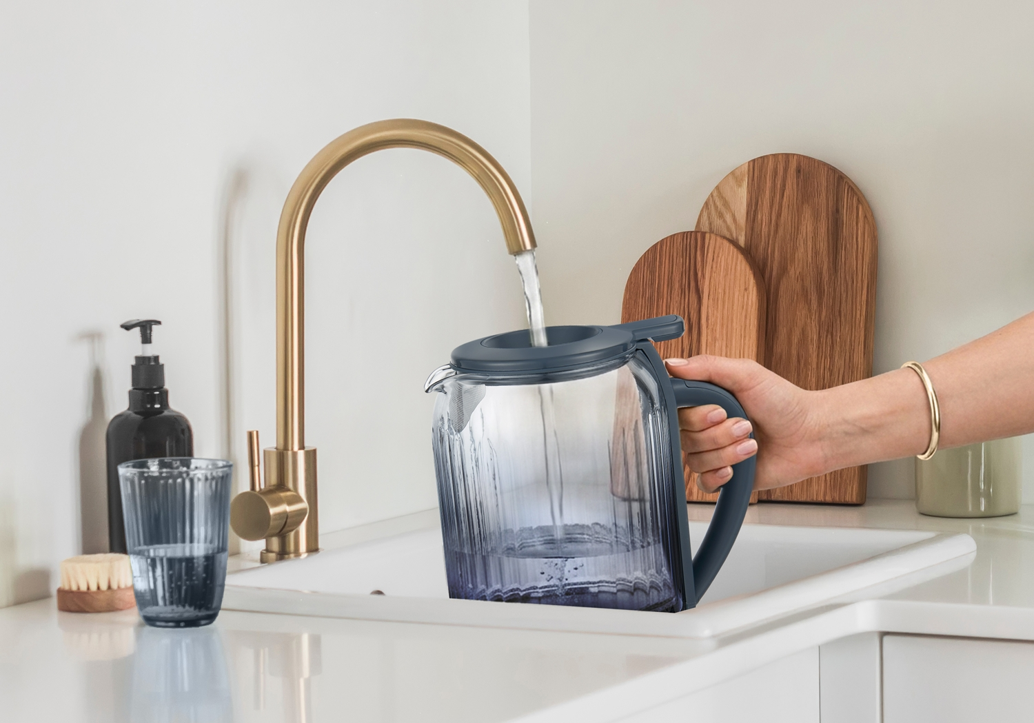 Una mano riempie una caraffa d'acqua in vetro con coperchio grigio sotto un rubinetto da cucina dorato.
