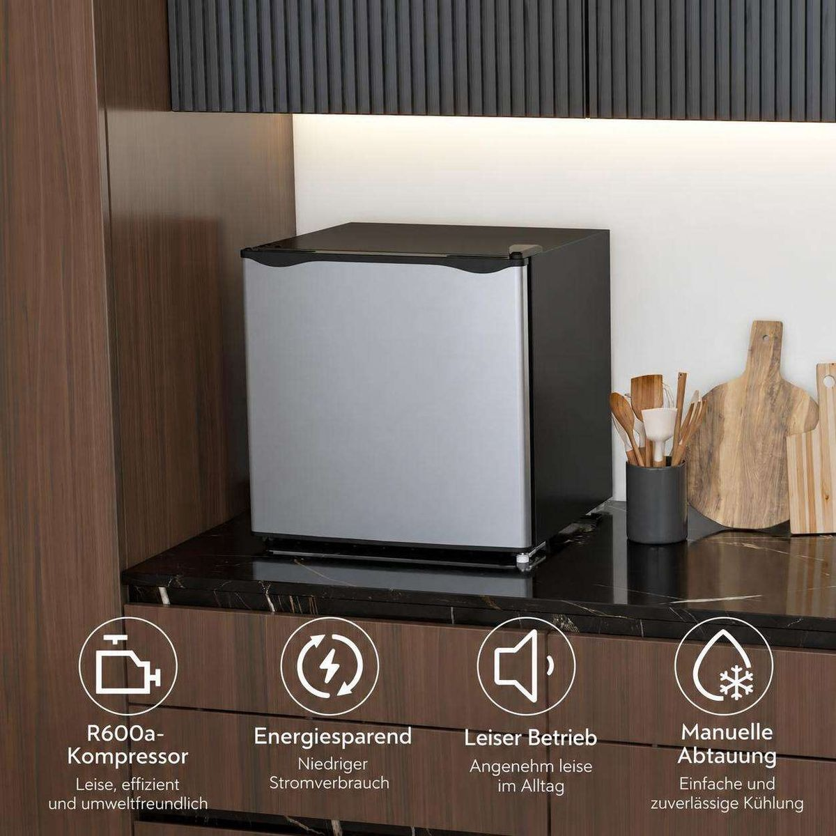 Ein kleiner silber-schwarzer Minikühlschrank steht auf dunkler Arbeitsplatte mit Utensilien und Icons.