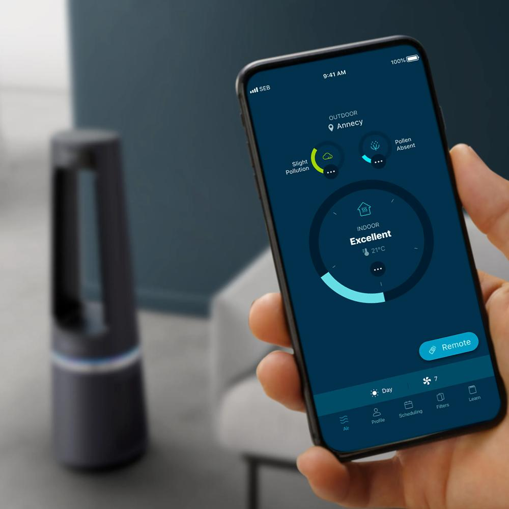 Hand hält Smartphone mit App-Bildschirm zur Luftqualitätsüberwachung, im Hintergrund ein passendes Luftreinigungsgerät.
