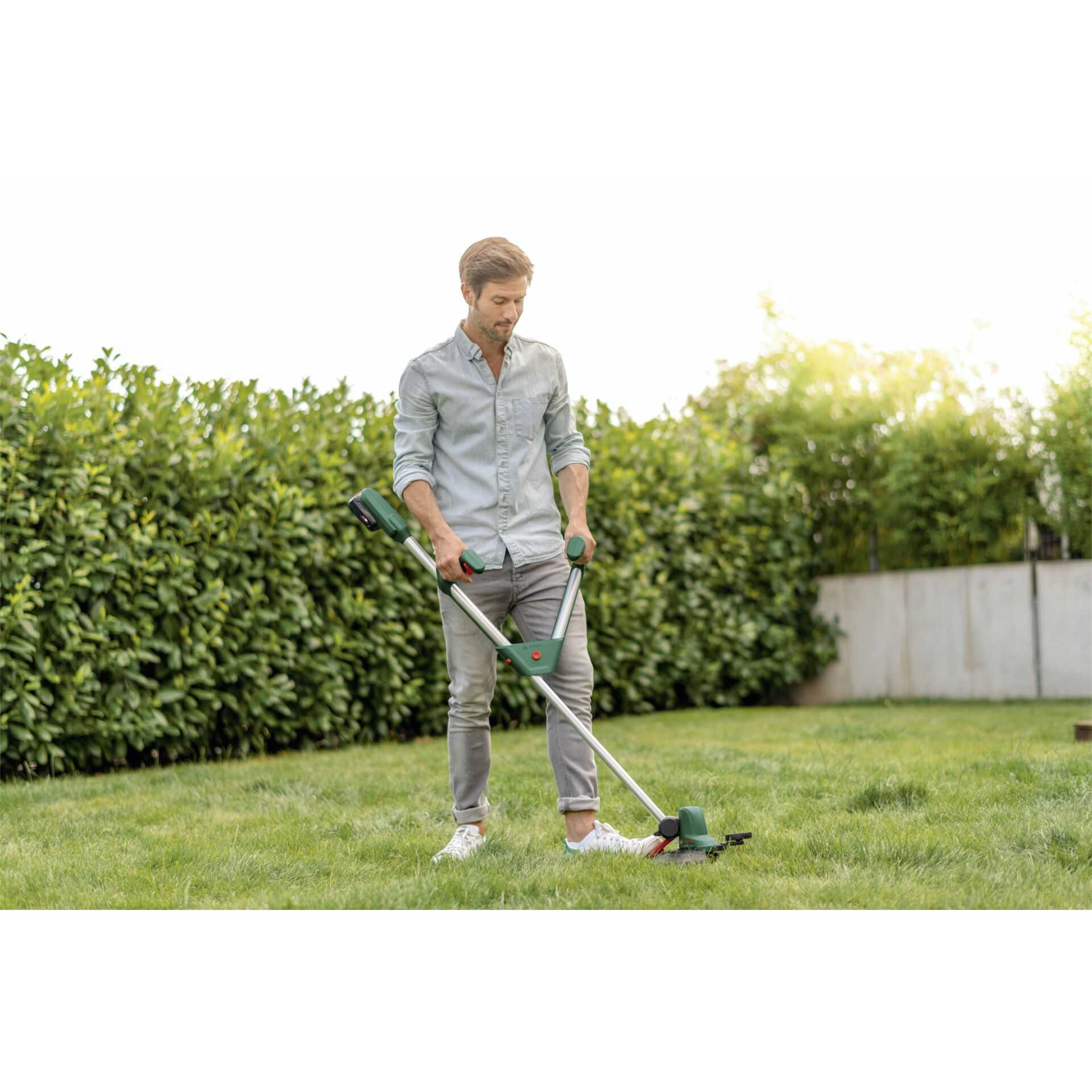 Ein Mann in hellem Hemd und Jeans trimmt im grünen Garten das Gras mit einem Akku-Rasentrimmer.