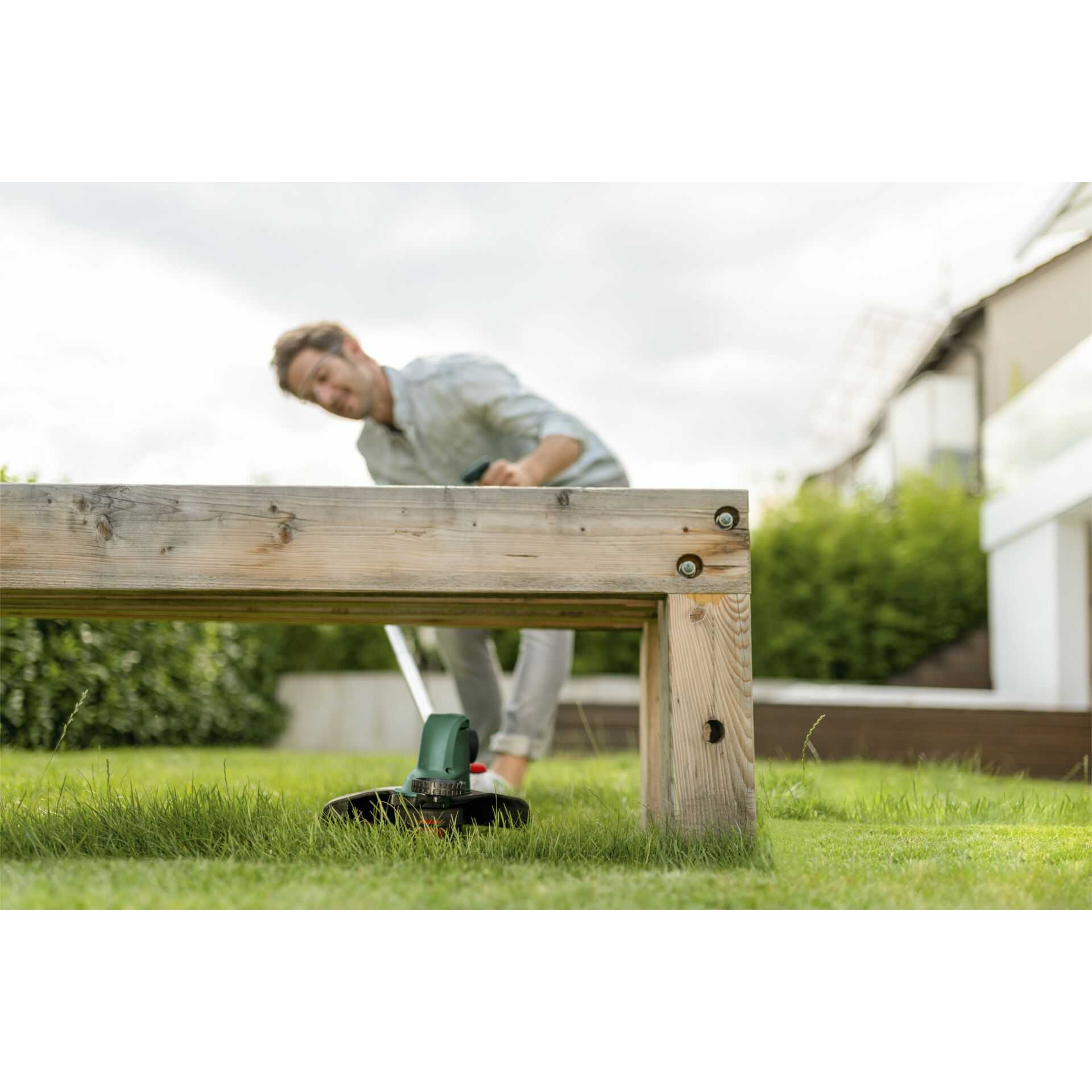 Ein Mann benutzt einen grünen Rasentrimmer in einem Garten in der Nähe einer Holzsbank.