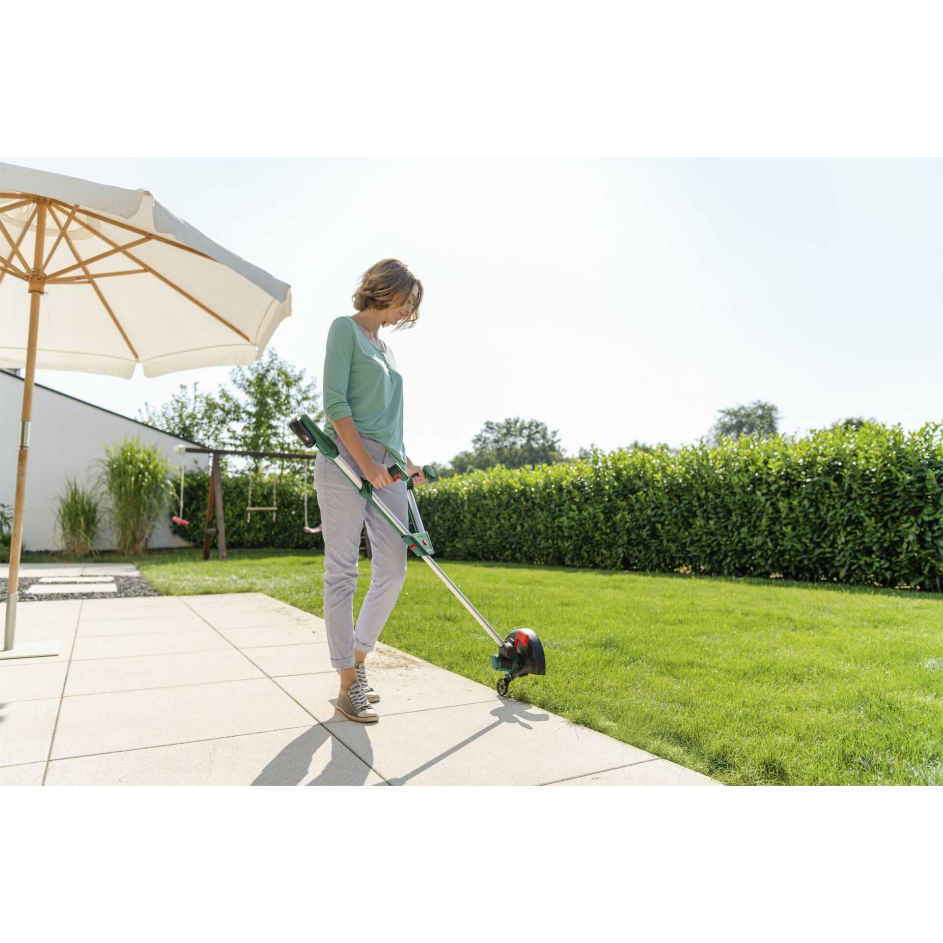 Eine Frau nutzt einen Rasentrimmer am Rand einer Steinterrasse im Garten.