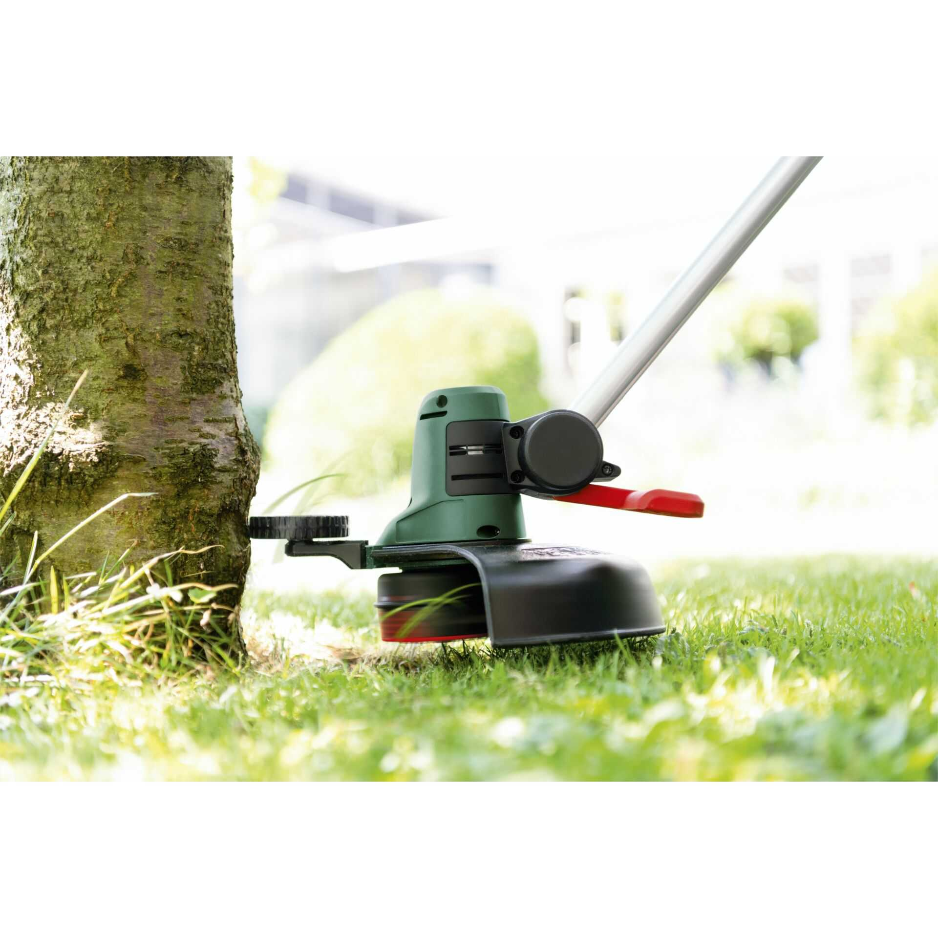Grüner Rasentrimmer schneidet Gras nah an einem Baumstamm in einem Garten.
