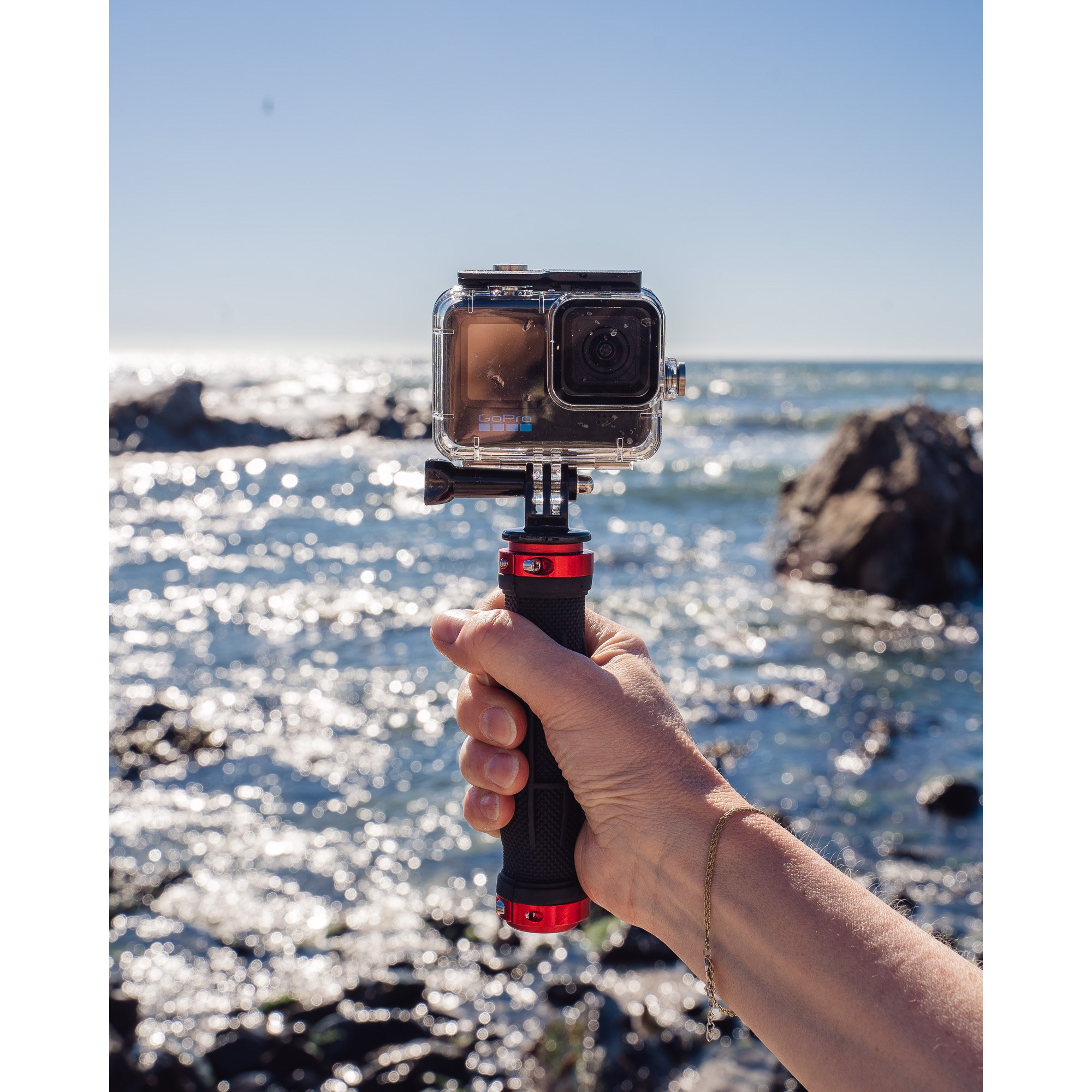 Een hand houdt een GoPro-camera in een waterdichte behuizing op een stok vast, met een glinsterende zee op de achtergrond.