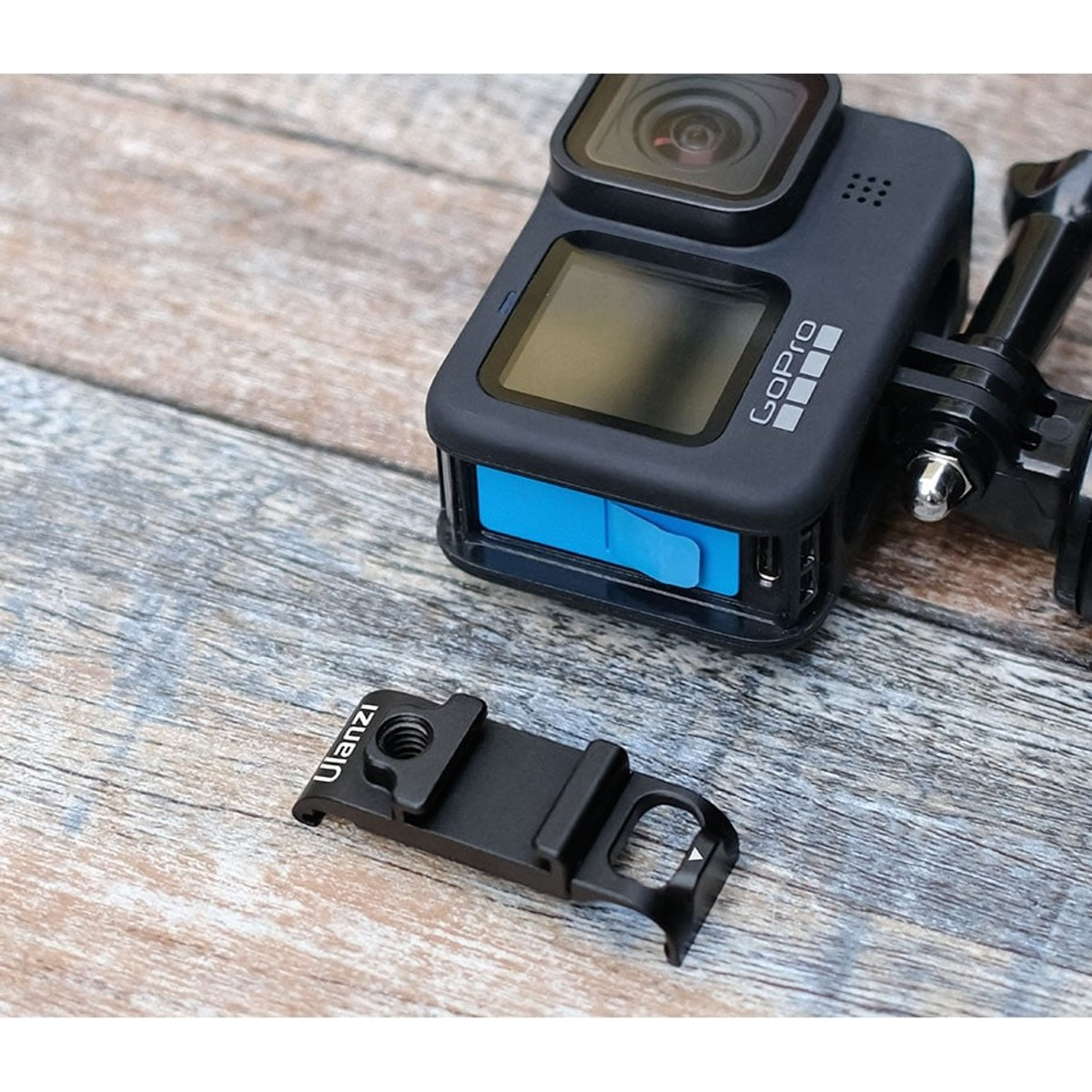 Een zwarte GoPro met blauwe accuklep ligt naast een metalen Ulanzi-bevestiging op hout.