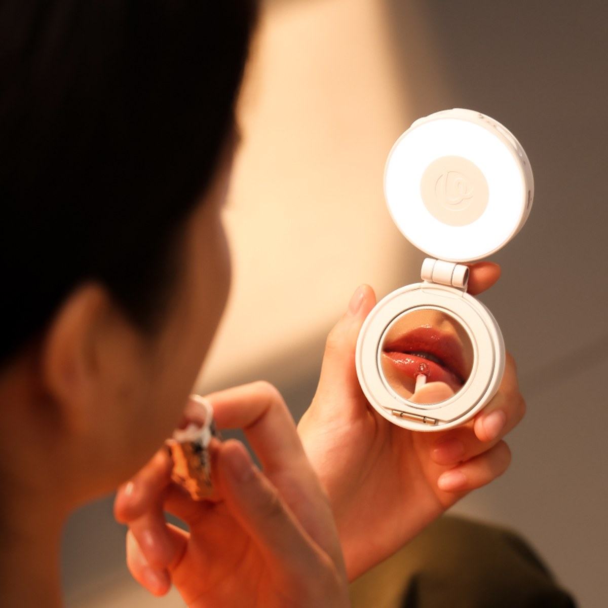 A person holds a small lit makeup mirror reflecting their lips while applying gloss.