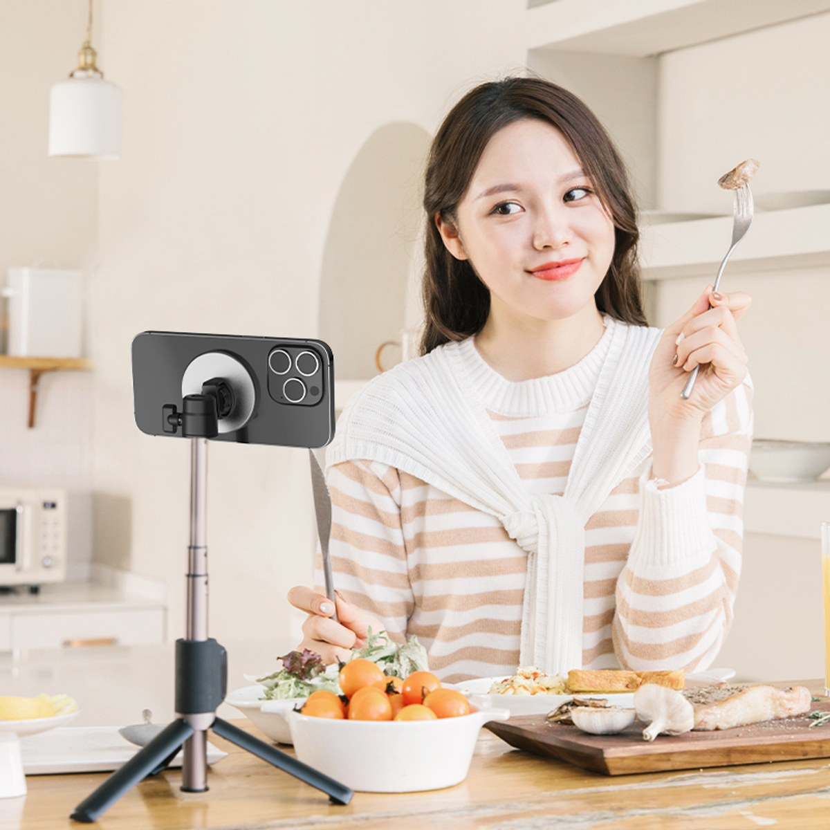Een vrouw in een wit-bruin gestreepte trui zit voor een telefoon op een statief en houdt een vork met eten vast.