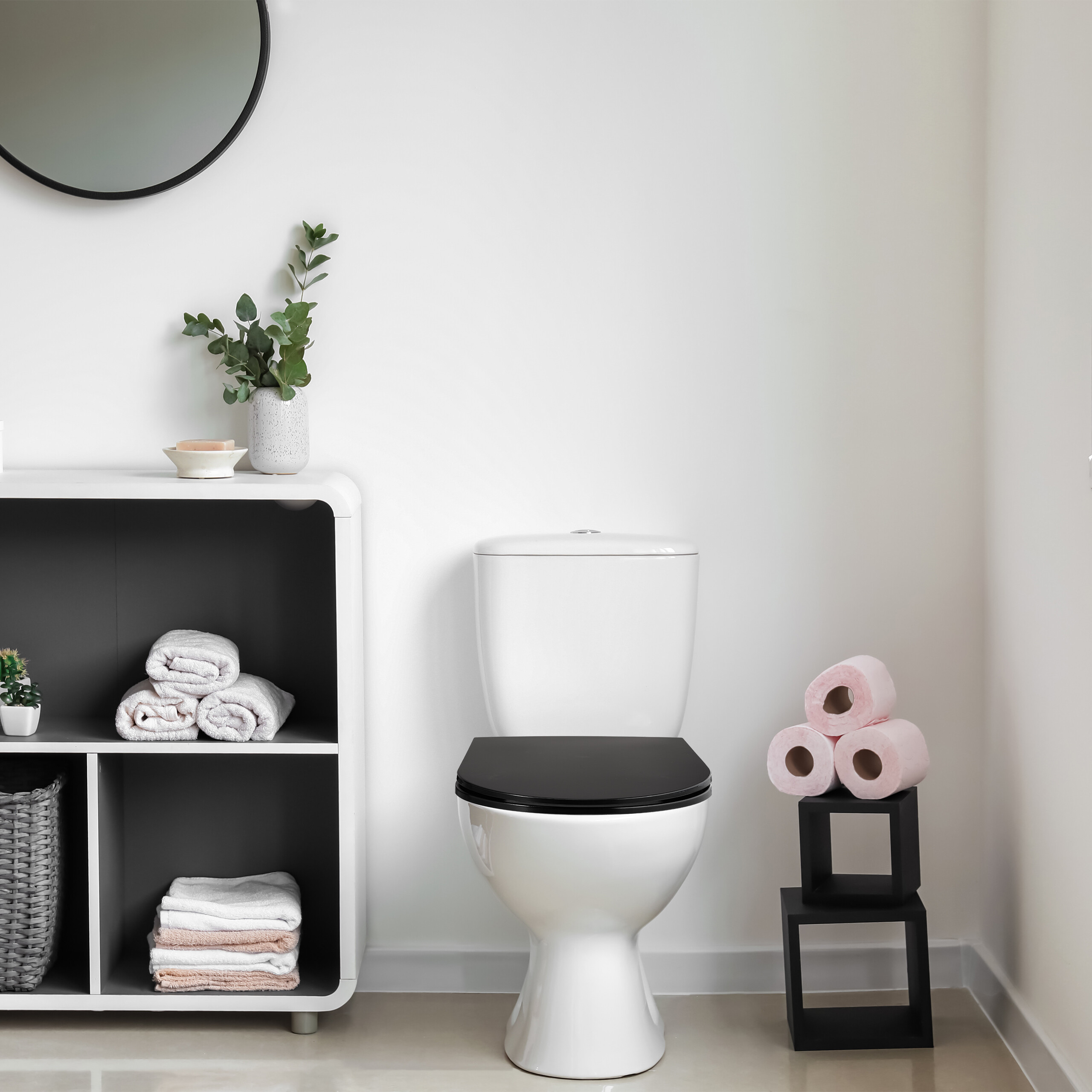 Moderne Toilette mit schwarzem Sitz, daneben ein weißes Regal mit Handtüchern und ein Turm aus rosa Toilettenpapier.