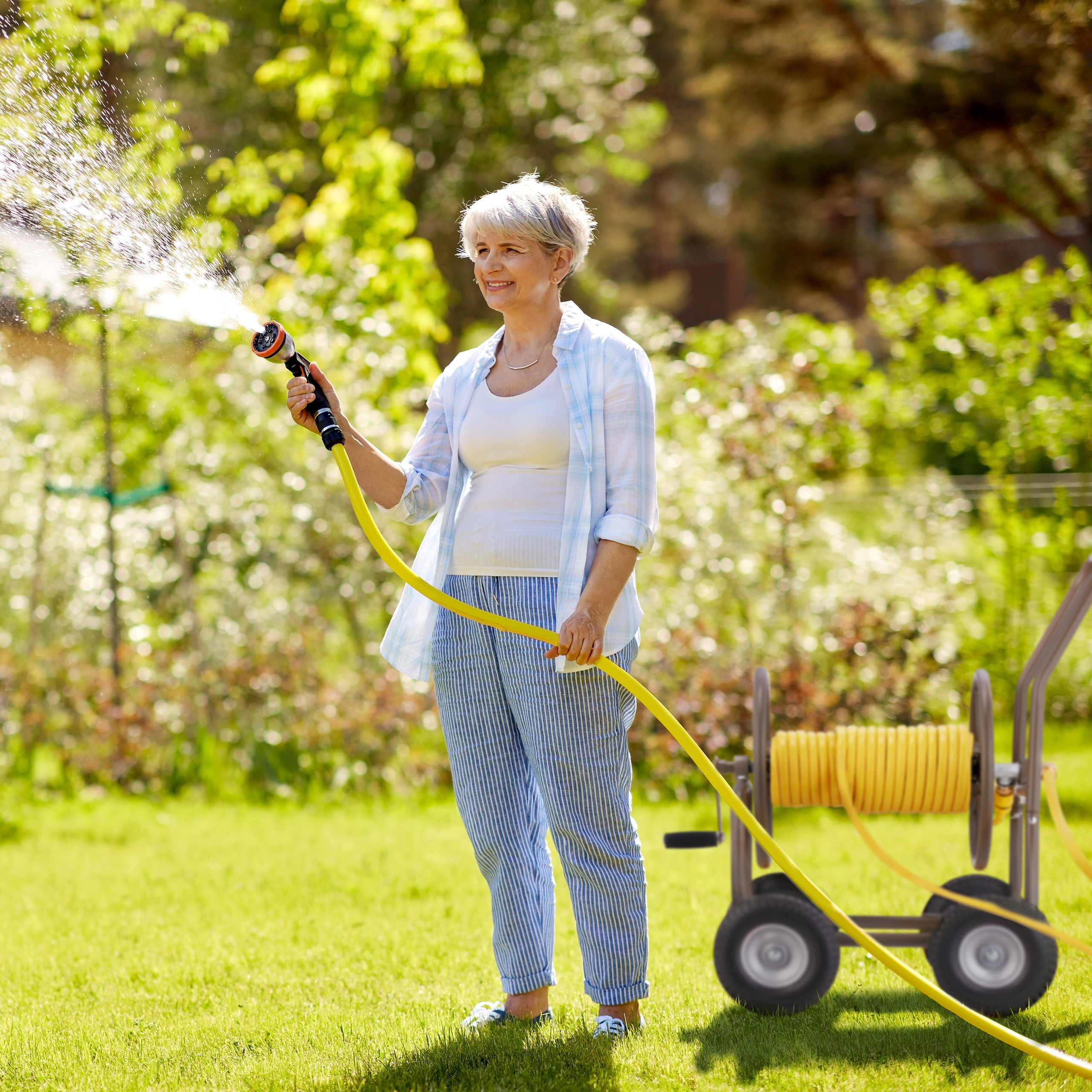 Ältere Frau gießt den Rasen im sonnigen Garten mit einem gelben Schlauch.