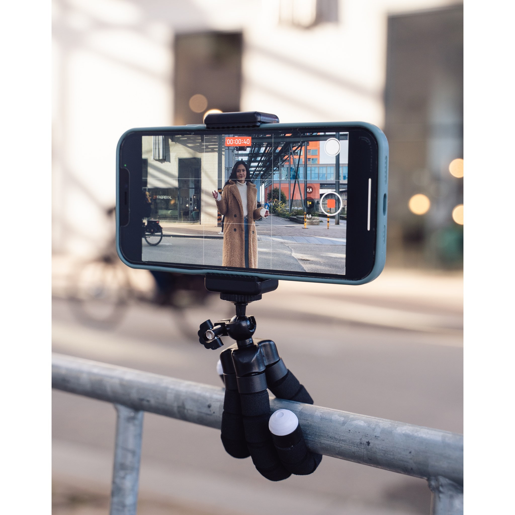 A smartphone on a small tripod records a woman in a brown coat on the street.