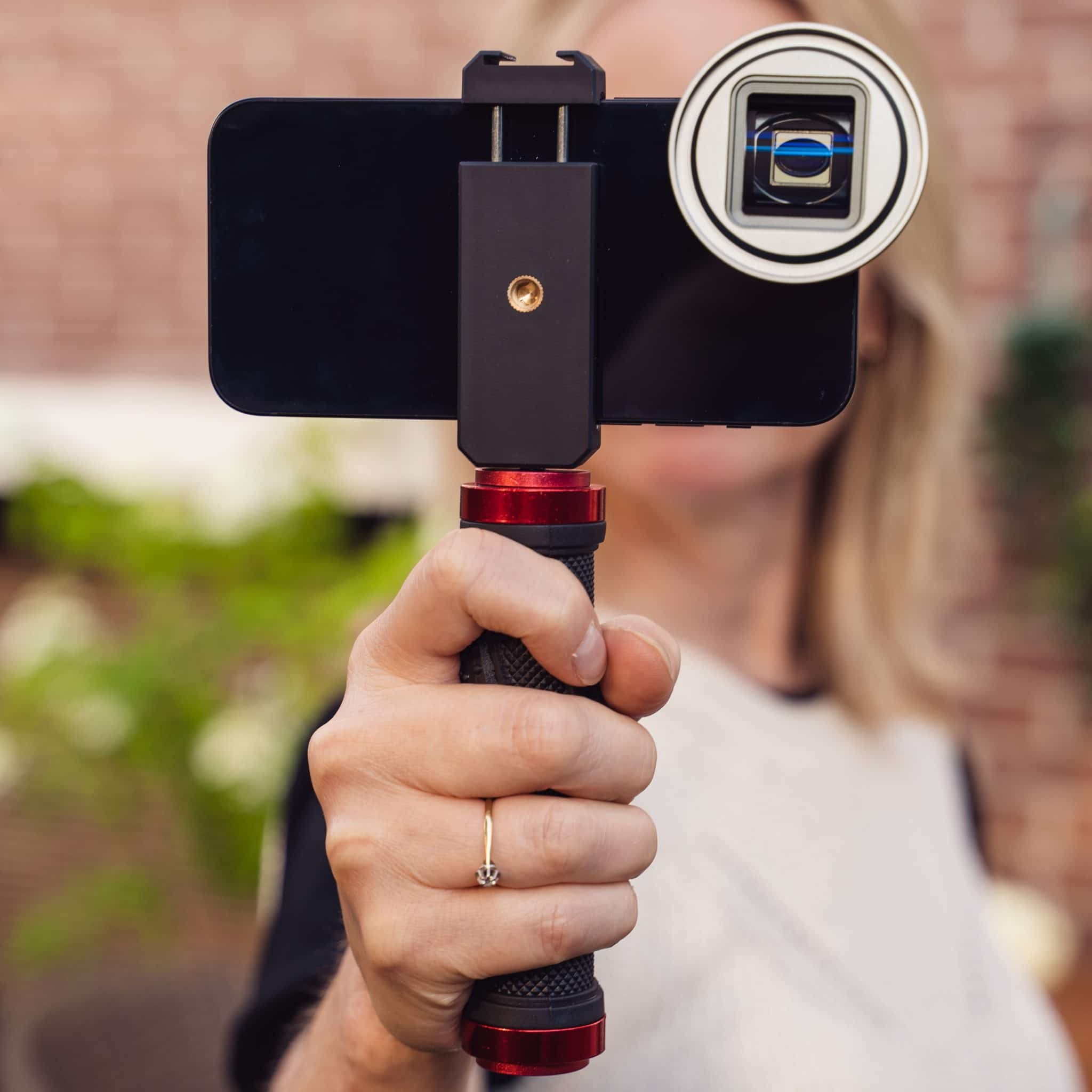 Eine Hand hält einen schwarzen Selfie-Stick mit Smartphone und aufgesetztem Zusatzobjektiv vor unscharfem Hintergrund.