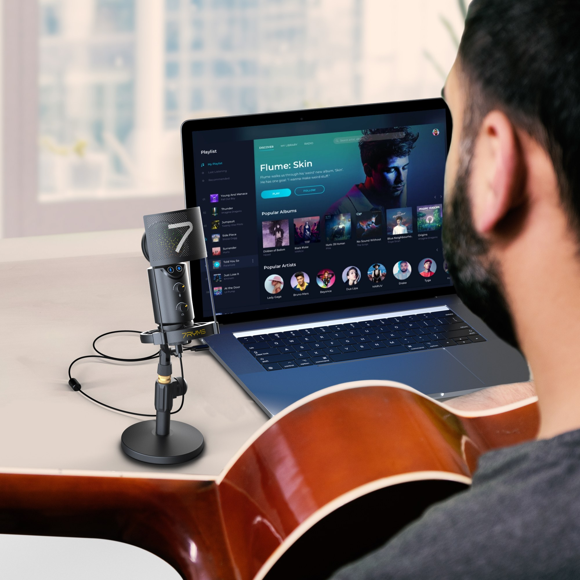 A musician sits before a laptop with music software, a microphone stands nearby and a guitar is in the foreground.