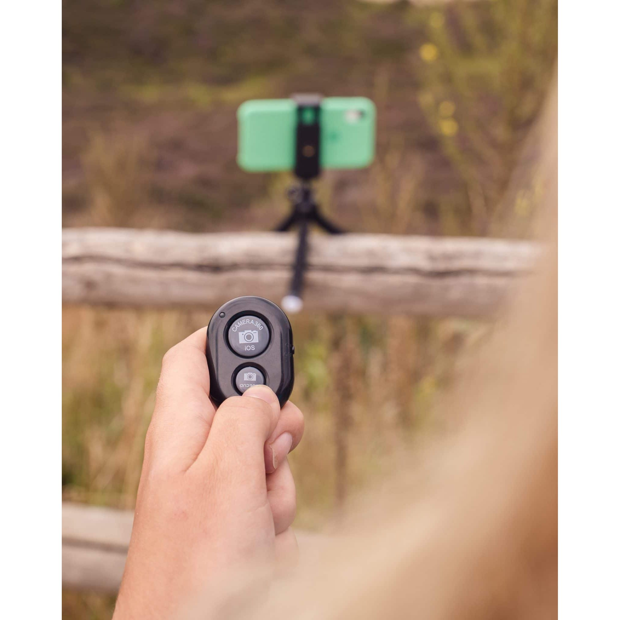 Hand hält eine Bluetooth-Fernbedienung vor einem auf einem Holzstab montierten Smartphone.