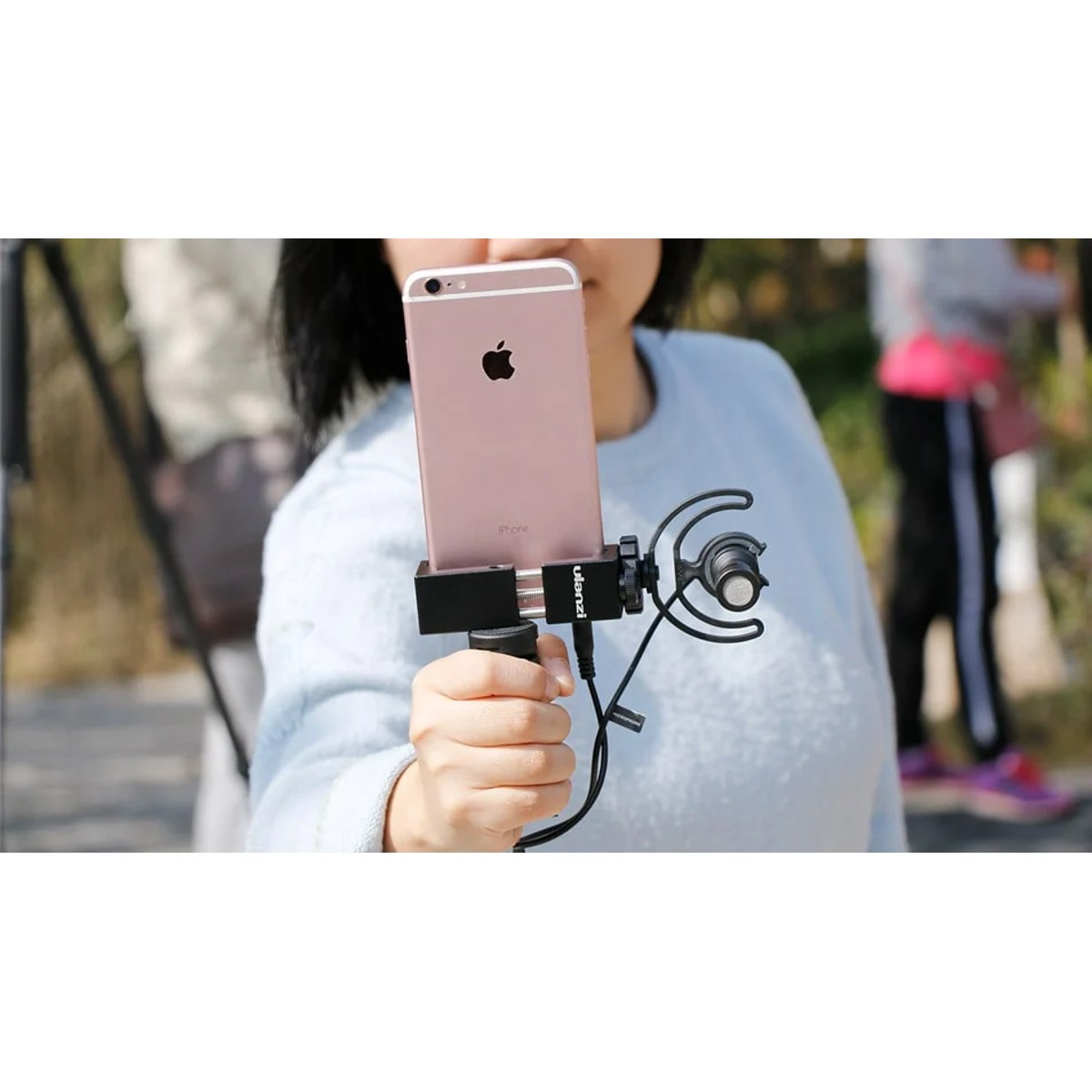 Eine Hand hält ein Smartphone in einer Halterung mit angeschlossenem Mikrofon, während eine Frau im Hintergrund steht.