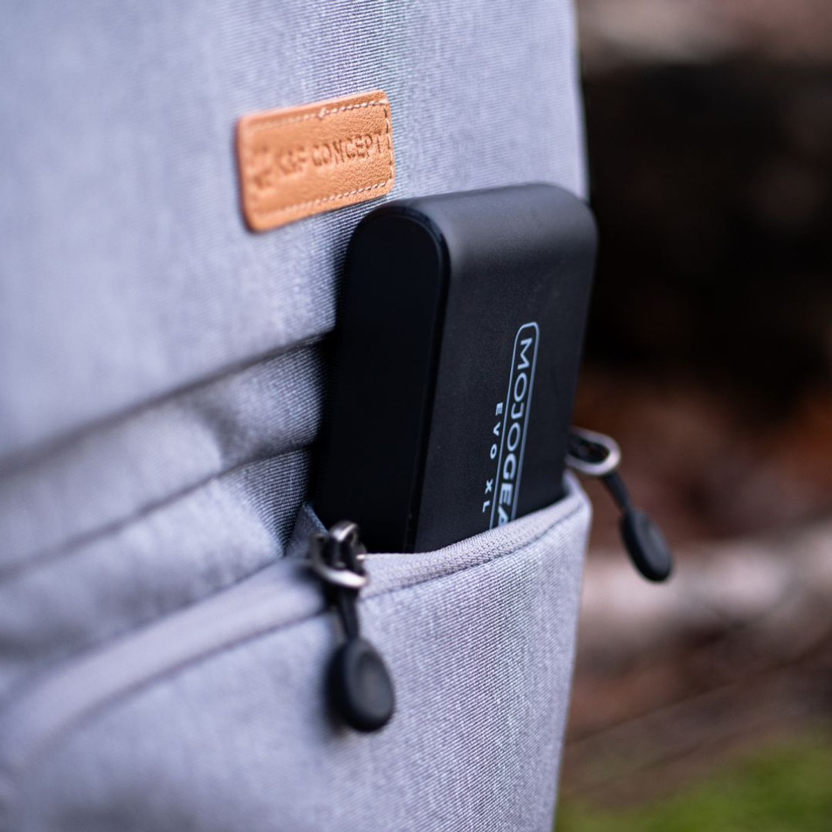 Une batterie externe Mojogear noire dans la poche avant d'un sac à dos gris avec logo en cuir marron.