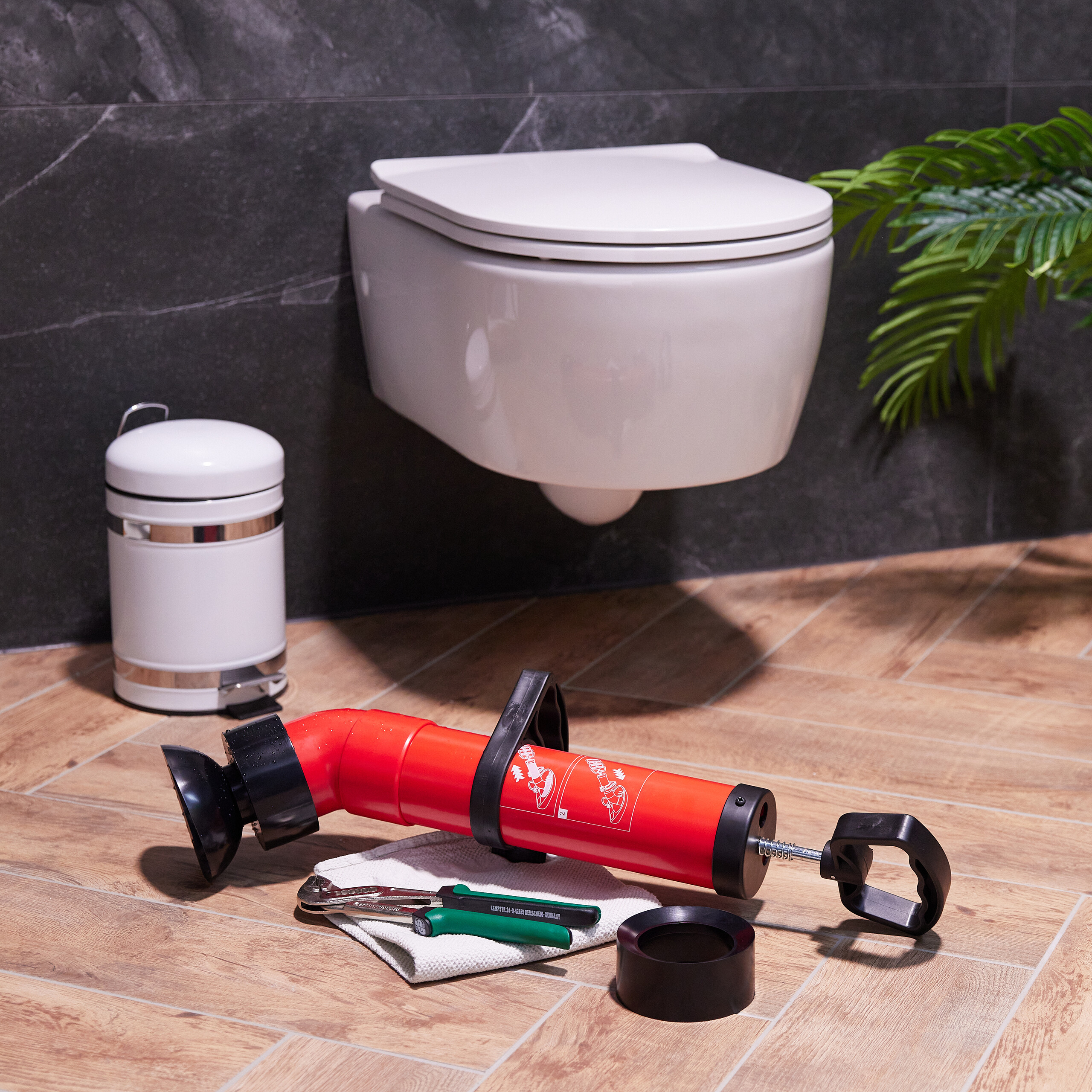 Ein roter WC-Entsorger liegt mit Werkzeugen auf dem Holzfußboden vor einer weißen hängenden Toilette und grauen Fliesen.