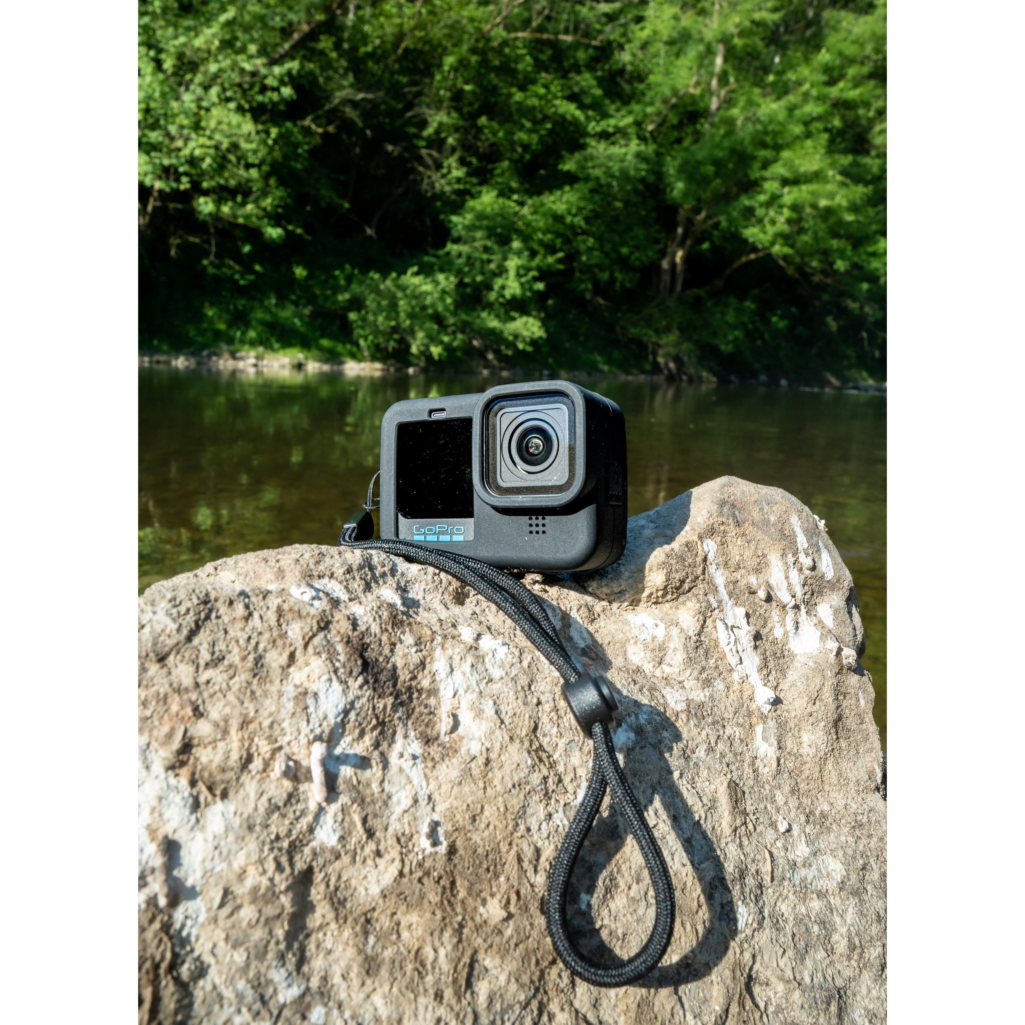 Een zwarte GoPro actiecamera met riem ligt op een grote rots bij een rivier, met groene bomen op de achtergrond.