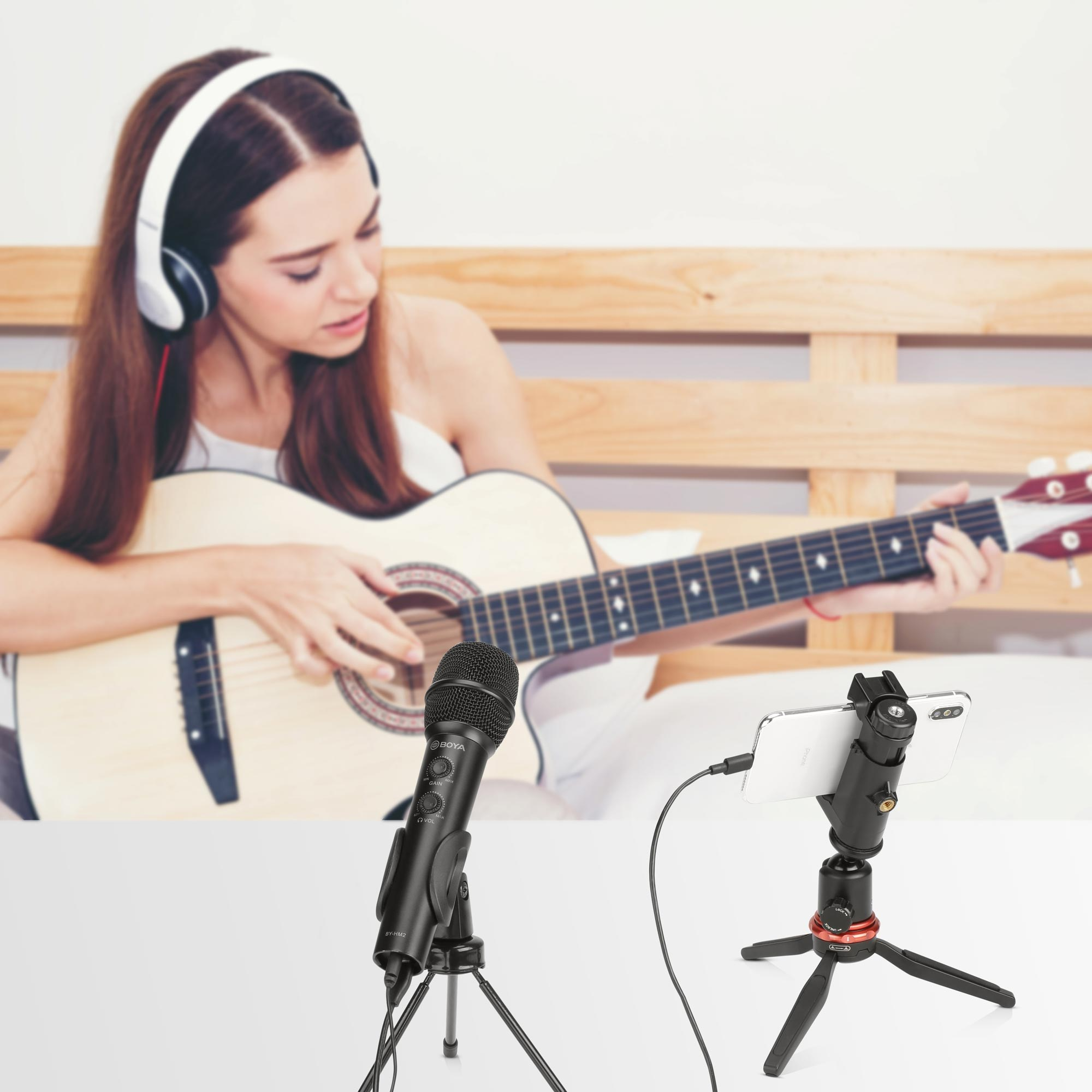 Ein schwarzes Mikrofon und ein Smartphone auf kleinen Stativen auf weißem Tisch, Hintergrund Frau mit Gitarre.