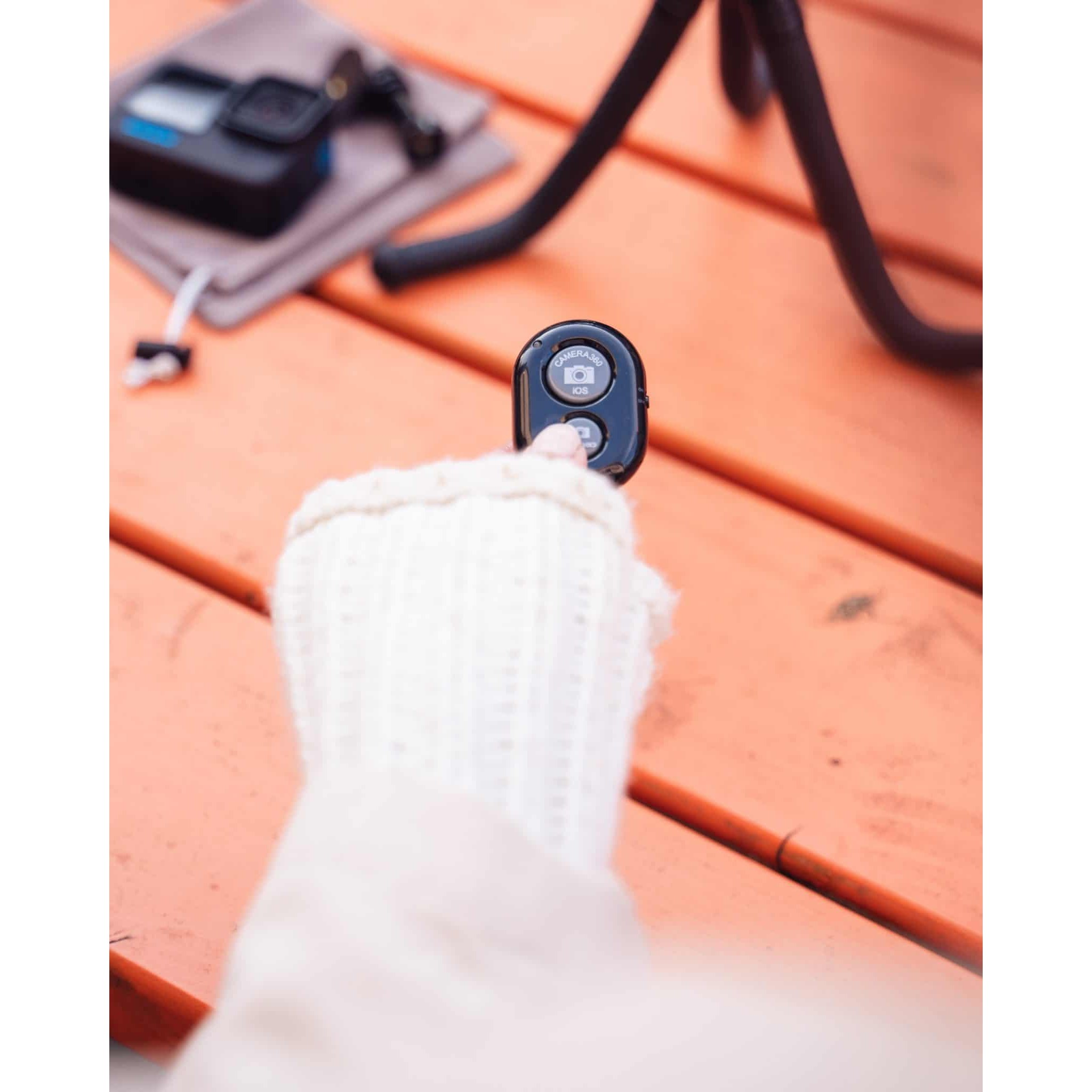 A hand in a white sweater holds a small black remote on an orange wooden table with a camera and tripod.