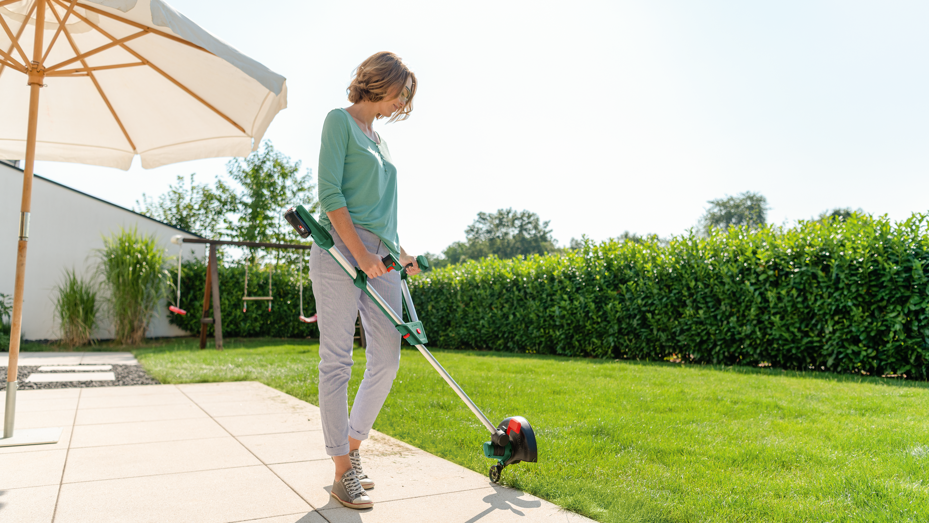 Eine Frau in grüner Bluse und heller Hose trimmt den Rasen im Garten mit einem Rasentrimmer.