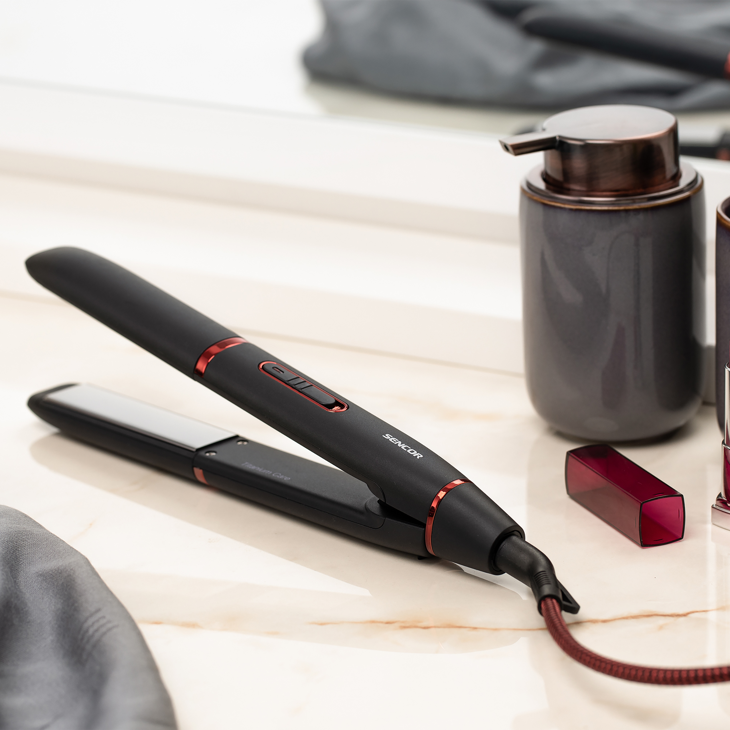 A black hair straightener with red accents on a marble table, next to a grey soap dispenser.