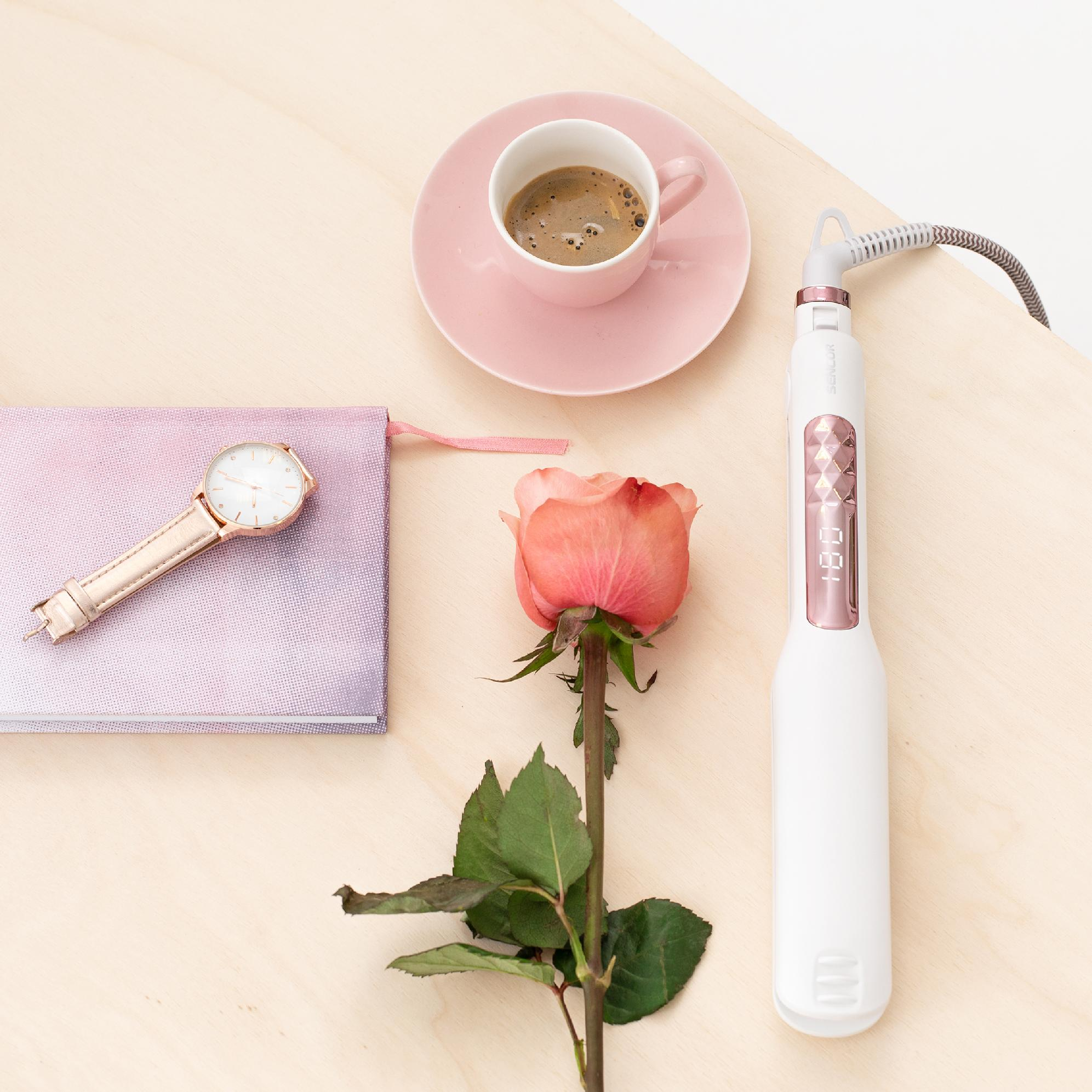 Una piastra per capelli bianca, tazza di caffè su piattino rosa, una rosa, un orologio e un taccuino sul legno.