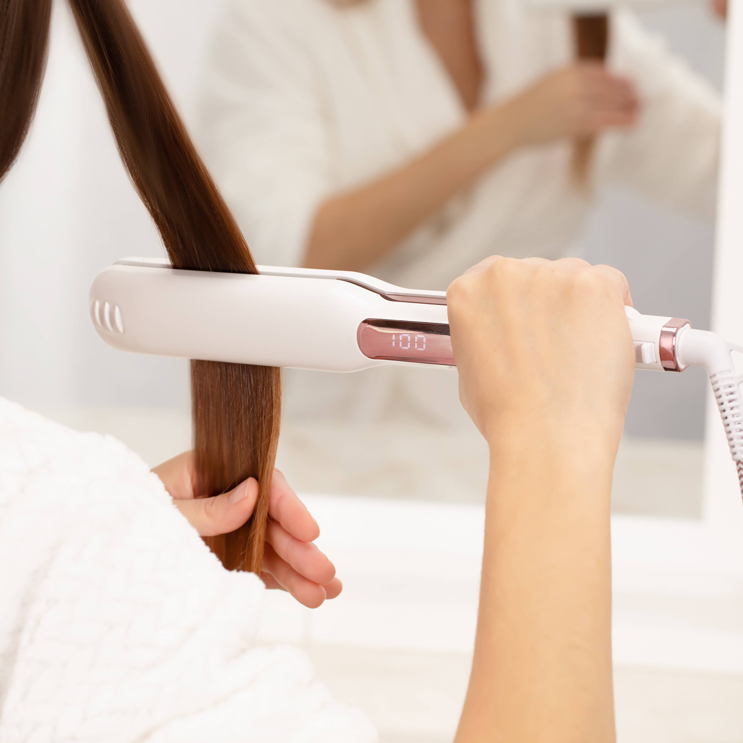 Una persona usa una piastra per capelli digitale bianca sui capelli castani davanti a uno specchio.