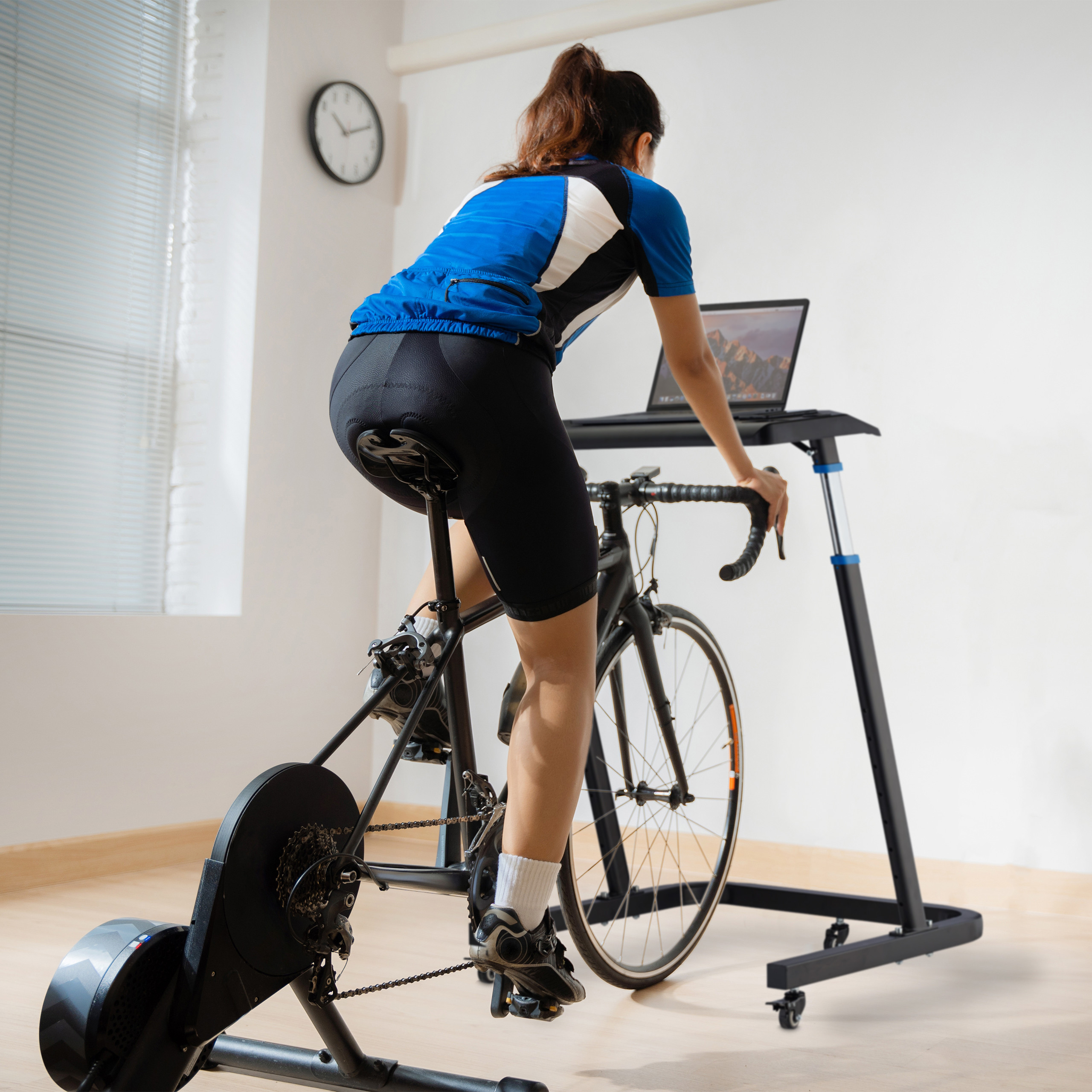 Eine Frau in blauer Sportkleidung radelt auf einem Heimtrainer vor einem Laptop in einem hellen Raum.