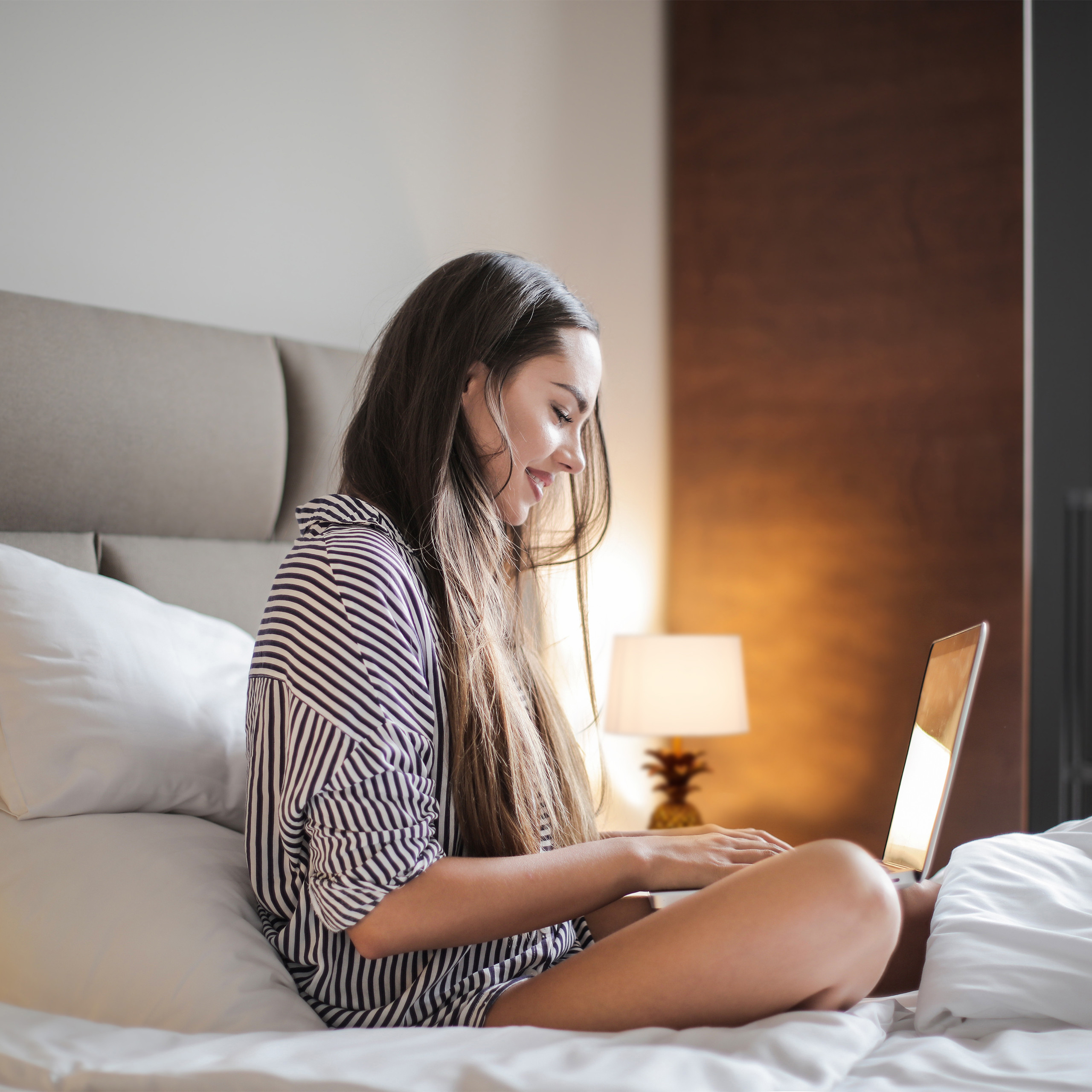 Eine Frau in gestreiftem Schlafanzug sitzt im Bett und arbeitet auf einem Laptop.
