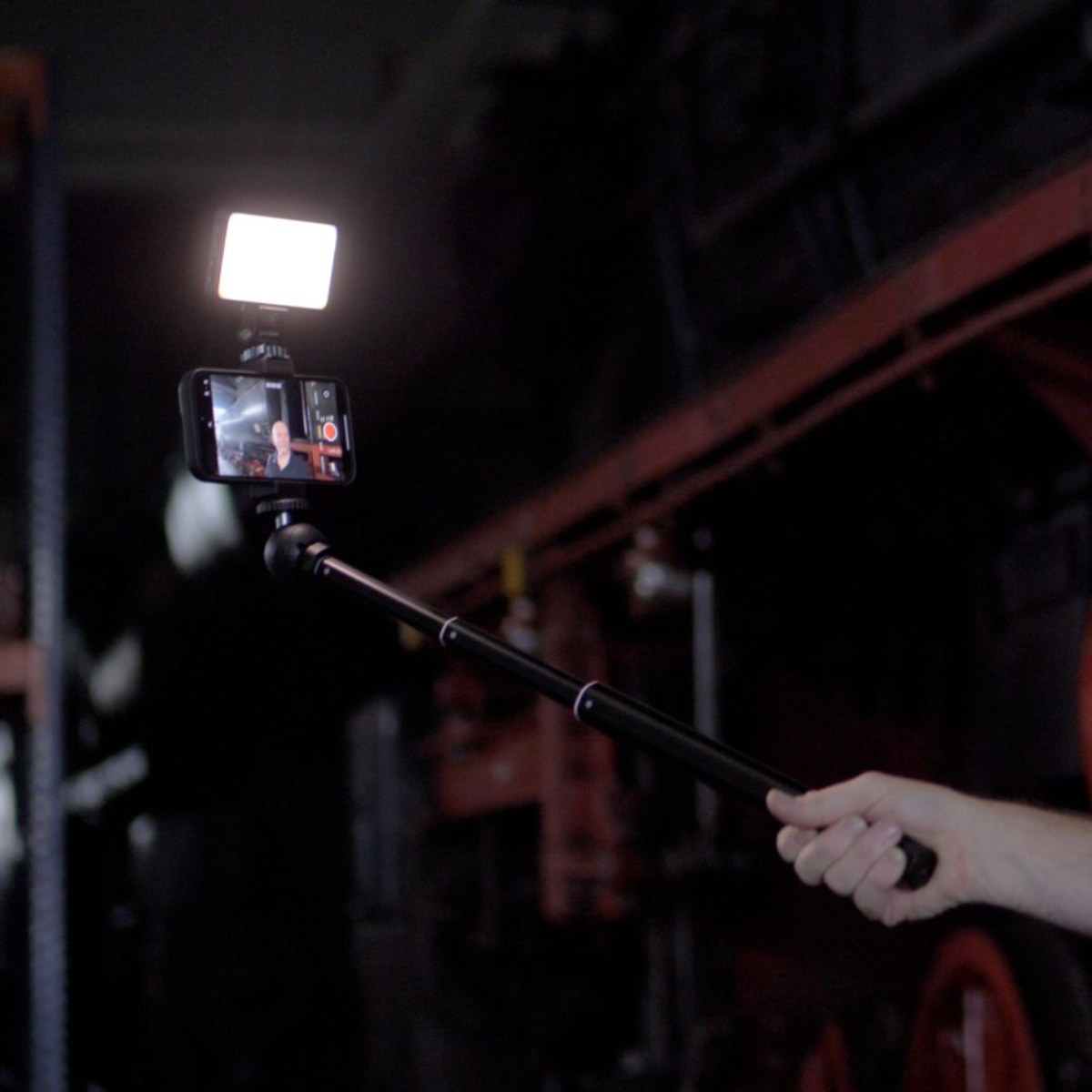 Hand holds selfie stick with smartphone and bright LED light in dark room.