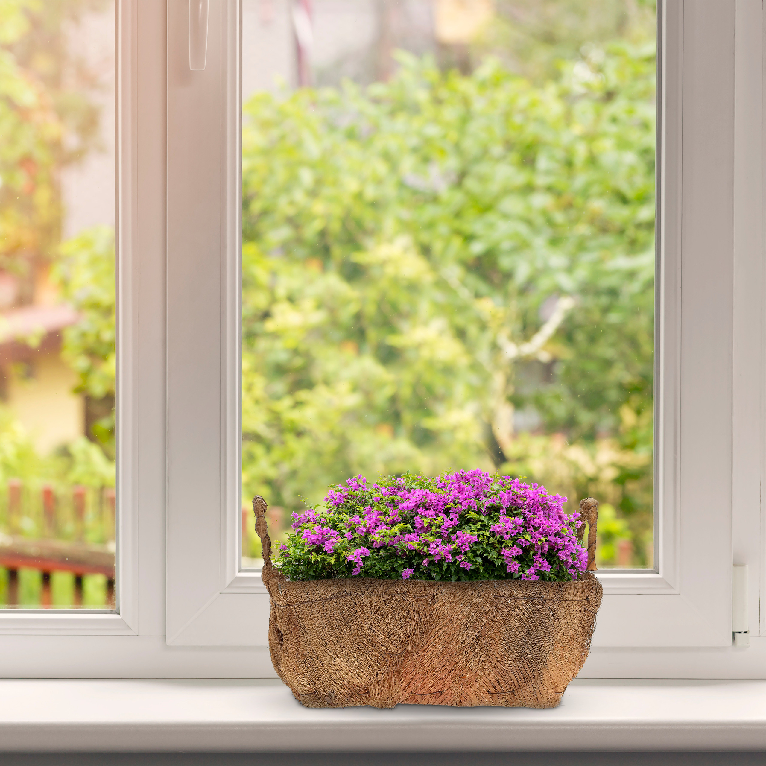 Ein Korb mit rosa Blumen steht auf einer weißen Fensterscheibe vor einem grünen Hintergrund.