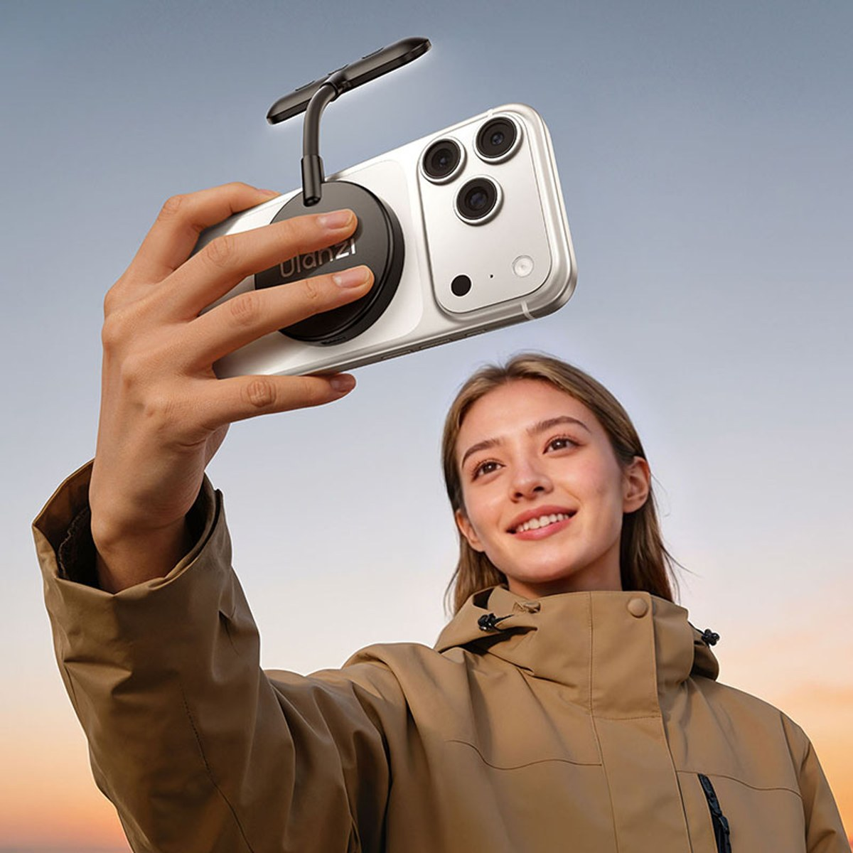 A woman in a beige coat takes a selfie with a smartphone attached to a small mount.