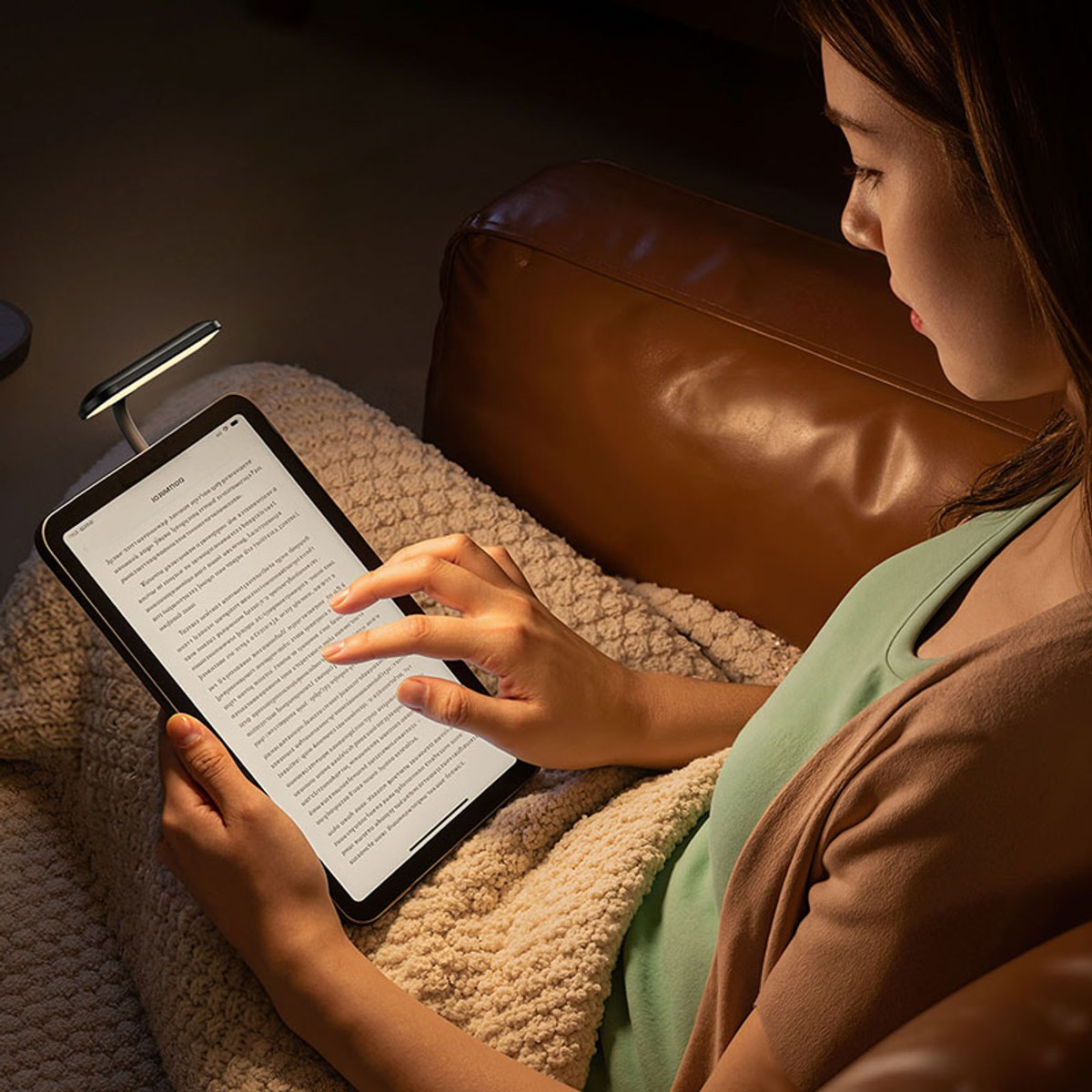 A woman sits on a leather couch at night, reading a tablet illuminated by a small clip-on reading light.