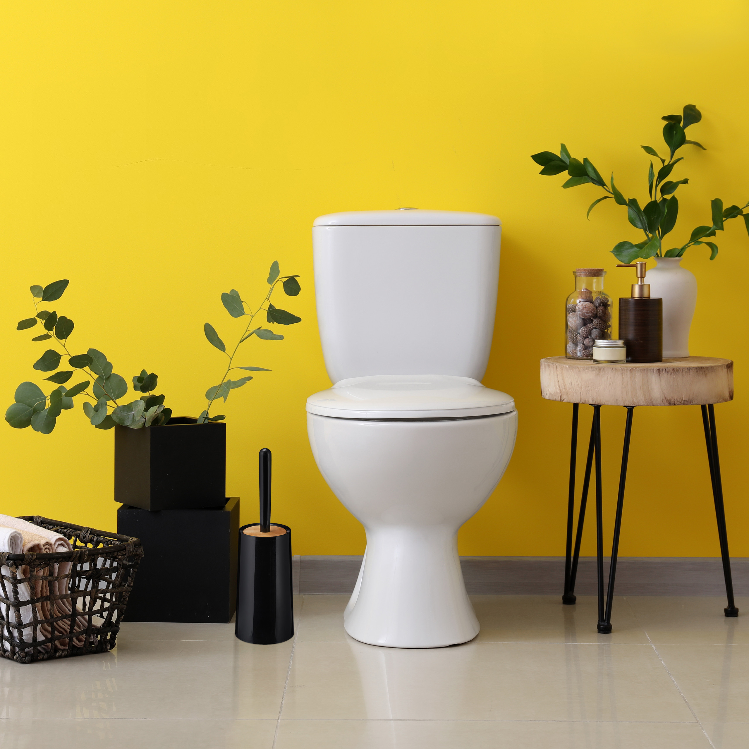 Eine weiße Toilette steht vor einer gelben Wand, flankiert von Pflanzen in schwarzen Töpfen und einem Beistelltisch.