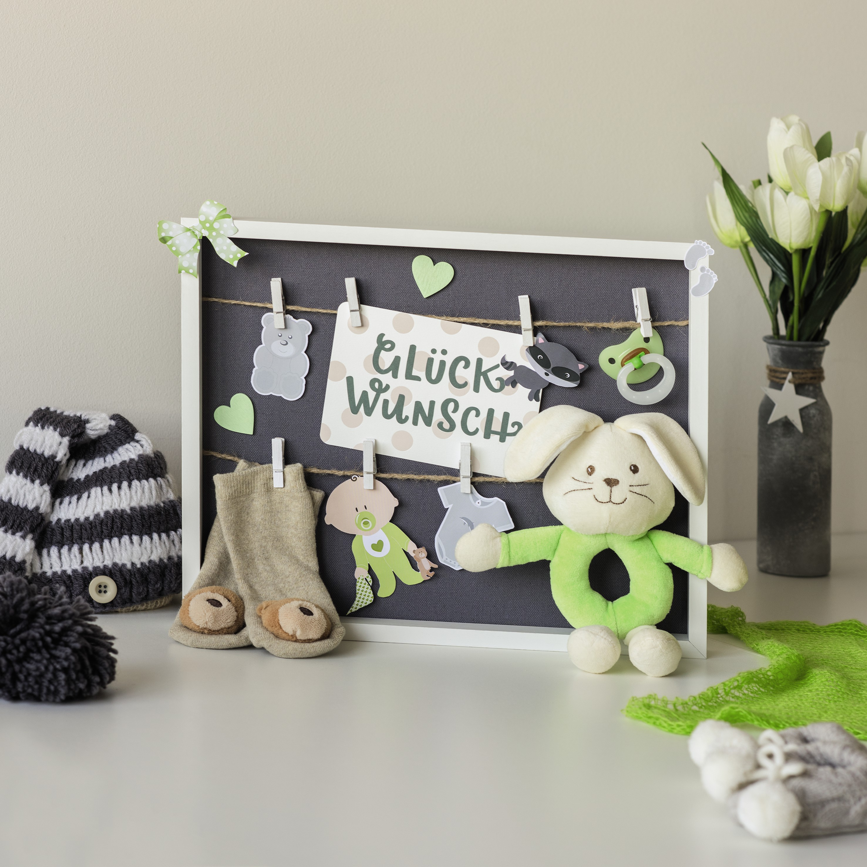 Tablica prezentowa na baby shower z napisem Glück Wunsch, pluszowym królikiem, skarpetkami i kwiatami na białej powierzchni.