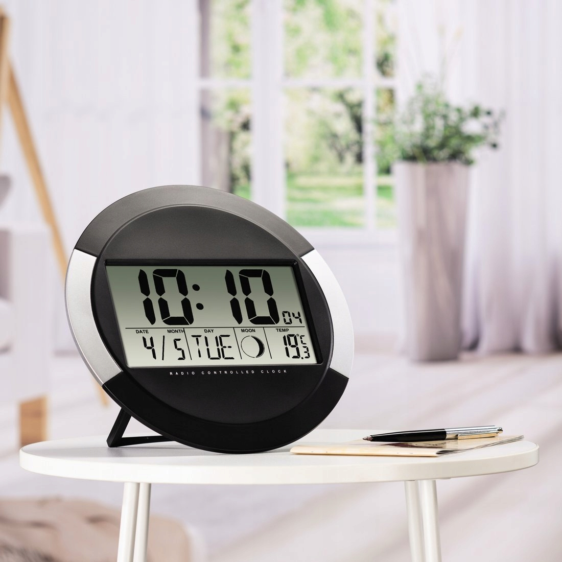 Eine schwarze und silberne Digitaluhr auf einem runden weißen Tisch, mit Notizbuch und Stift daneben in einem Raum.
