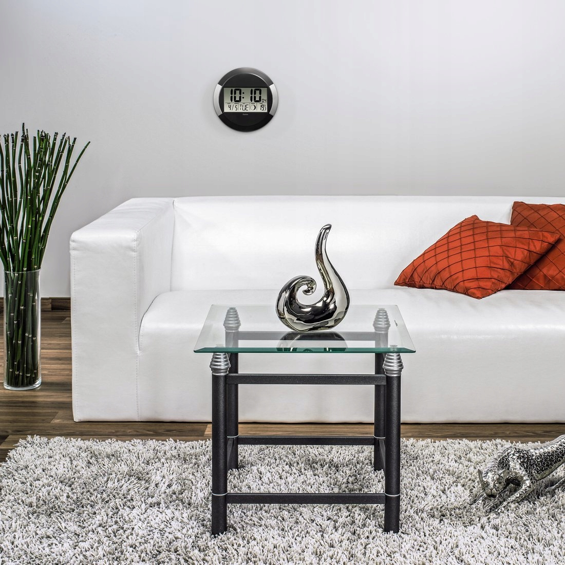 Wohnzimmer mit weißem Sofa, Glastisch, silberner Skulptur, digitaler Wanduhr und hohen grünen Bambusrohren.