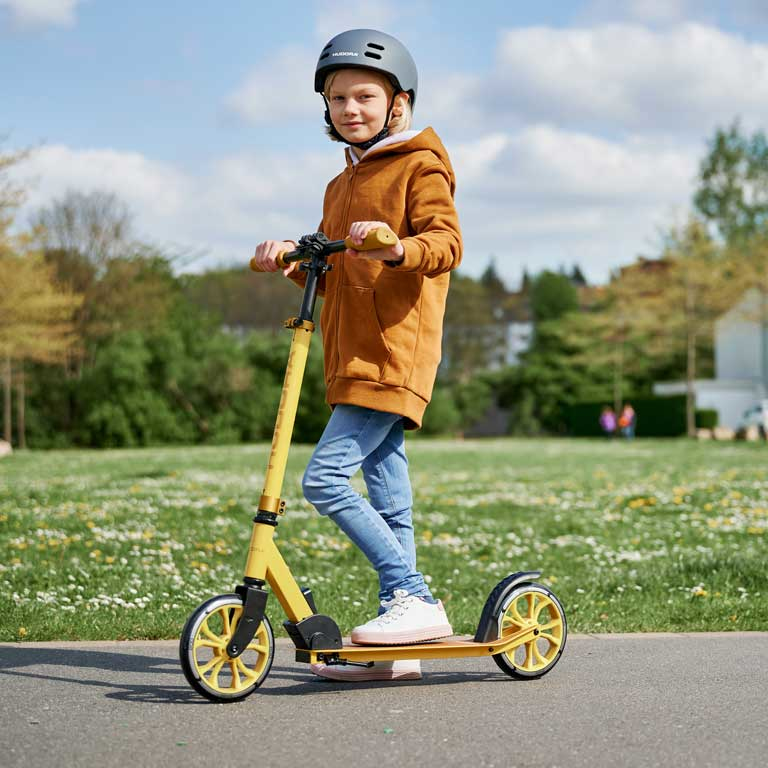 Ein Kind mit Helm und gelbem Hoodie fährt auf einem gelben Tretroller auf einem Parkweg.