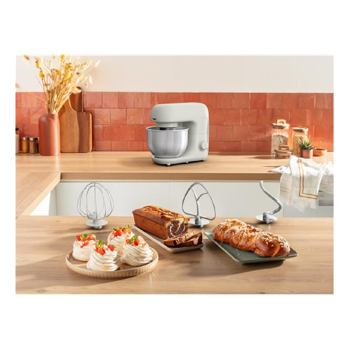 Un piano cucina con un'impastatrice bianca, accessori, dolci, pane e prodotti da forno sullo sfondo.